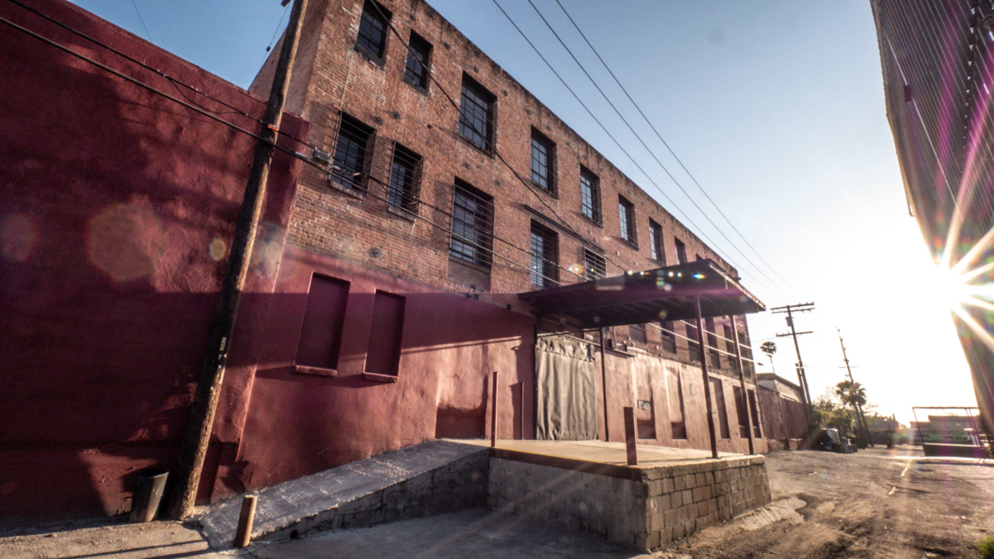 The image shows a red brick building with multiple windows, a loading dock, and a gravel path, illuminated by sunlight in a narrow alley.