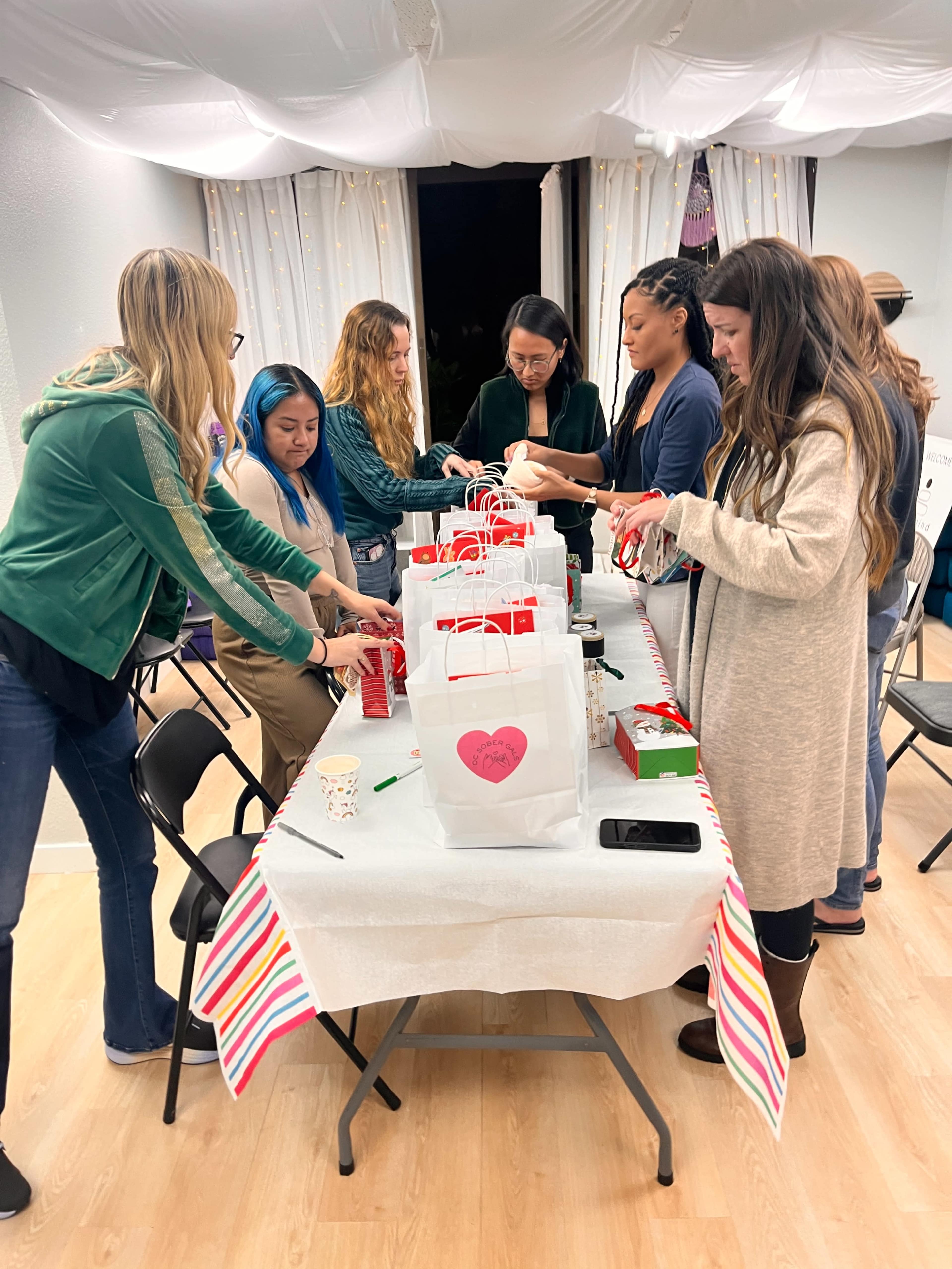A group of women are assembling gift bags at a table decorated with a striped tablecloth in a well-lit indoor space.