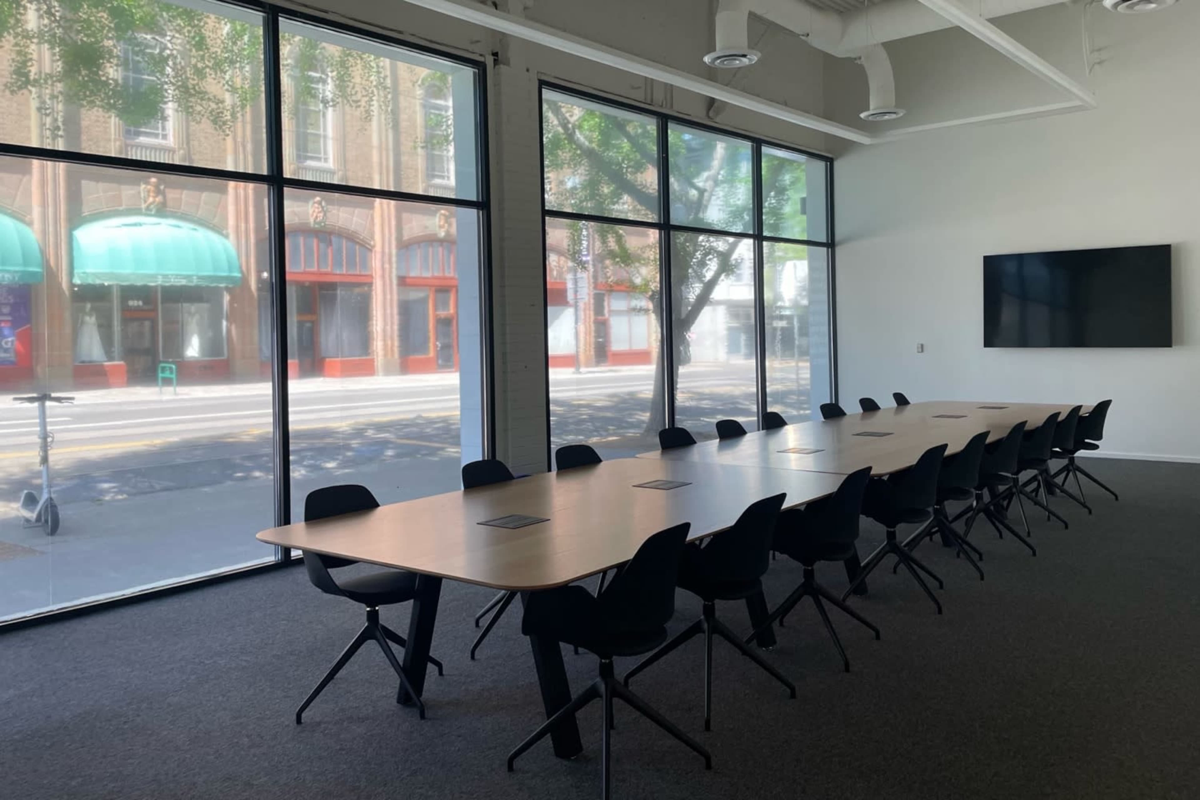 A long conference table with black chairs is positioned in a bright, minimally furnished room featuring large windows overlooking a street with storefronts.