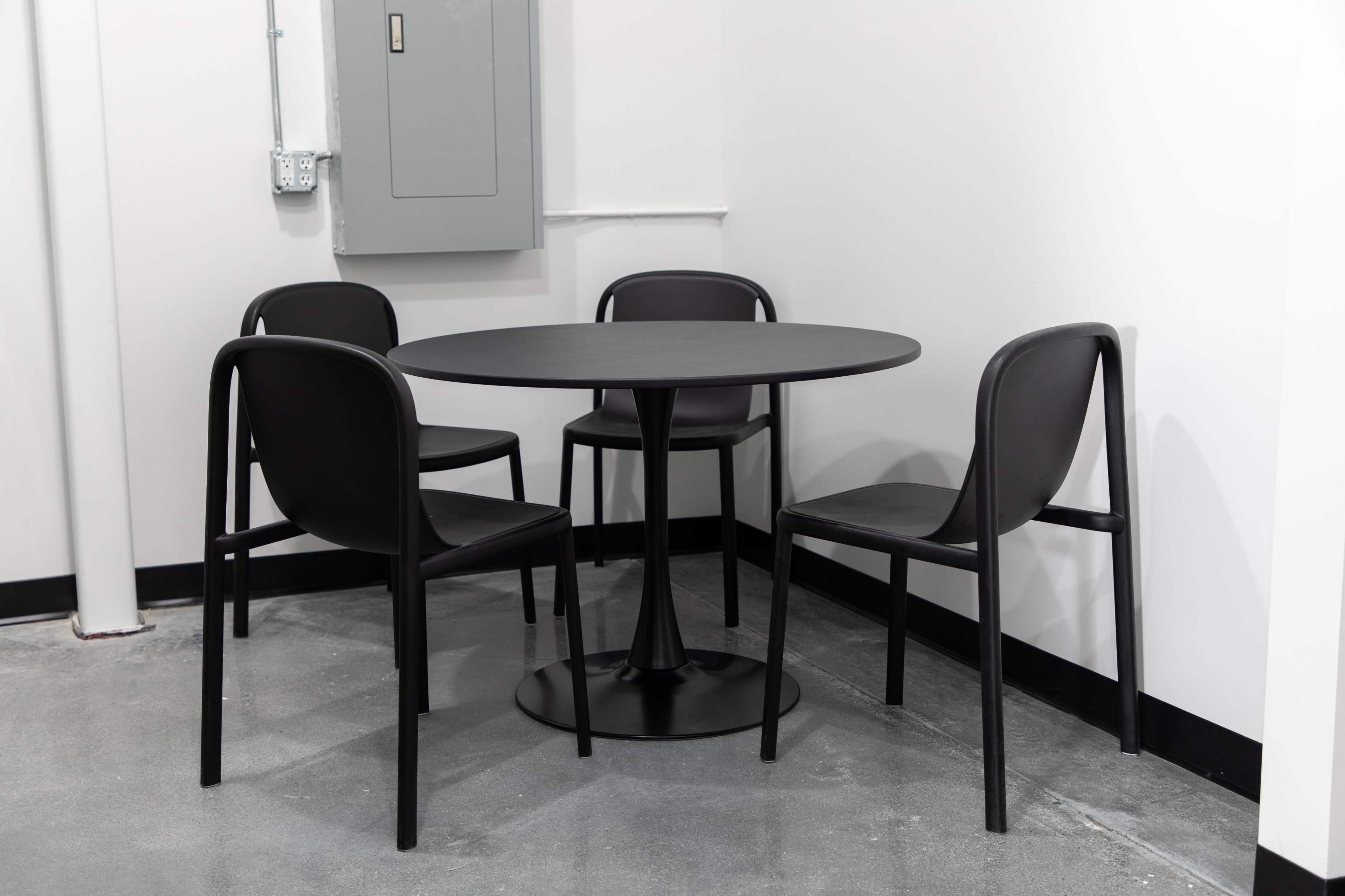 A round table with four black chairs is placed in a corner of a room with grey flooring and white walls.