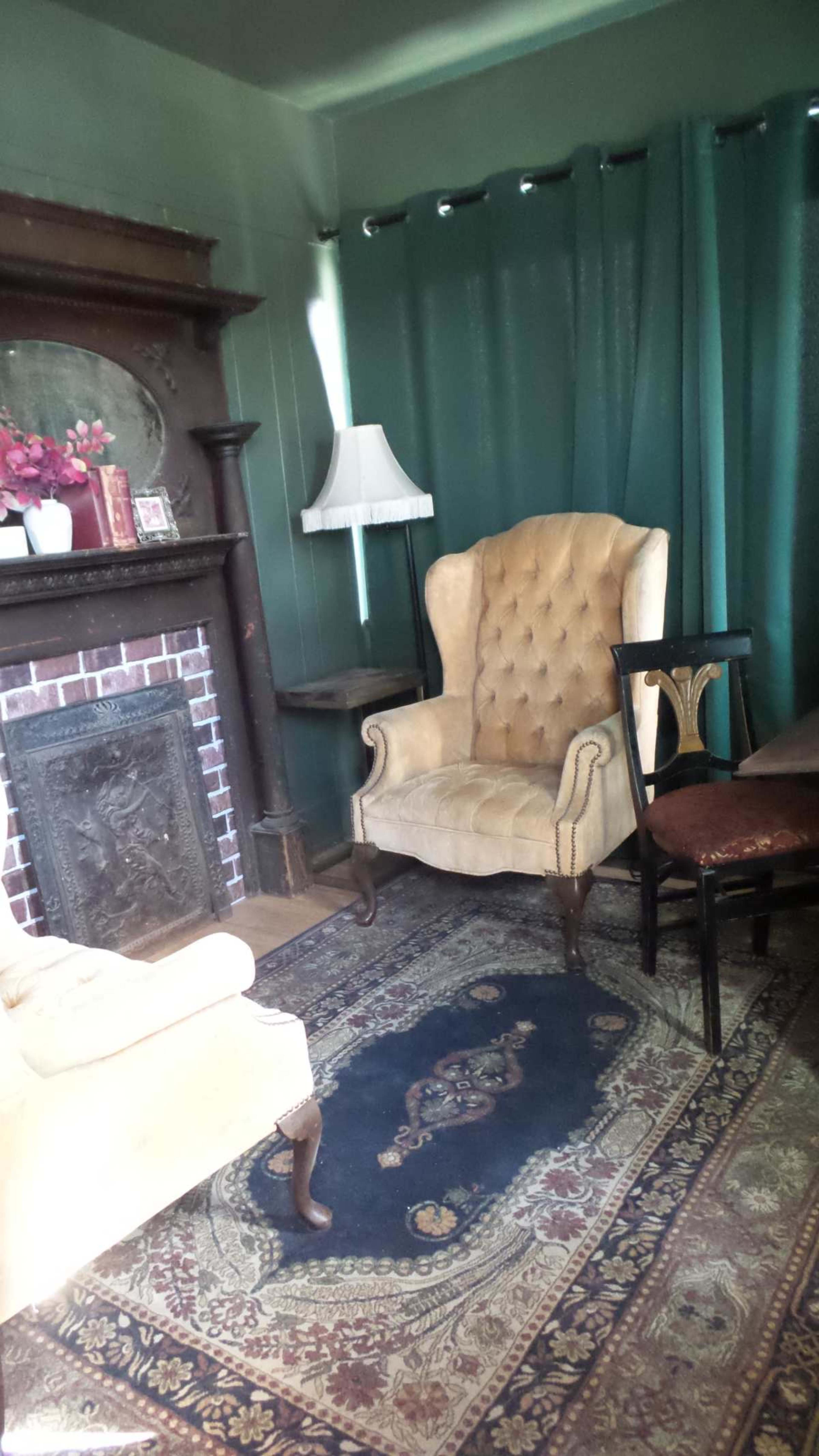 A living room with a beige wingback chair, a small wooden side table, and a fireplace surrounded by a patterned rug and green curtains.