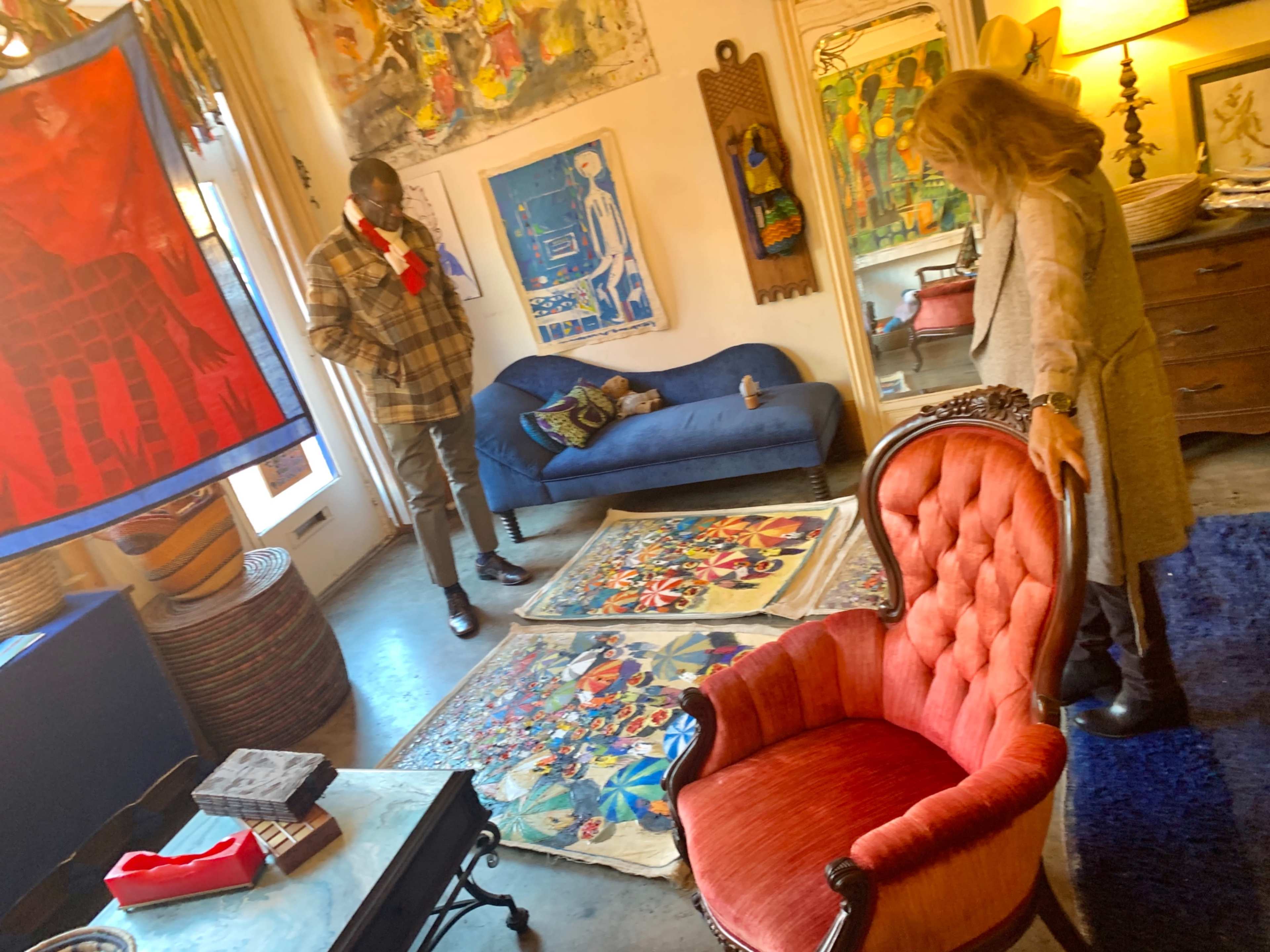 A man in a plaid coat and a woman in a long sweater stand in a room decorated with colorful artwork and patterned rugs, with a vintage orange chair in the foreground.