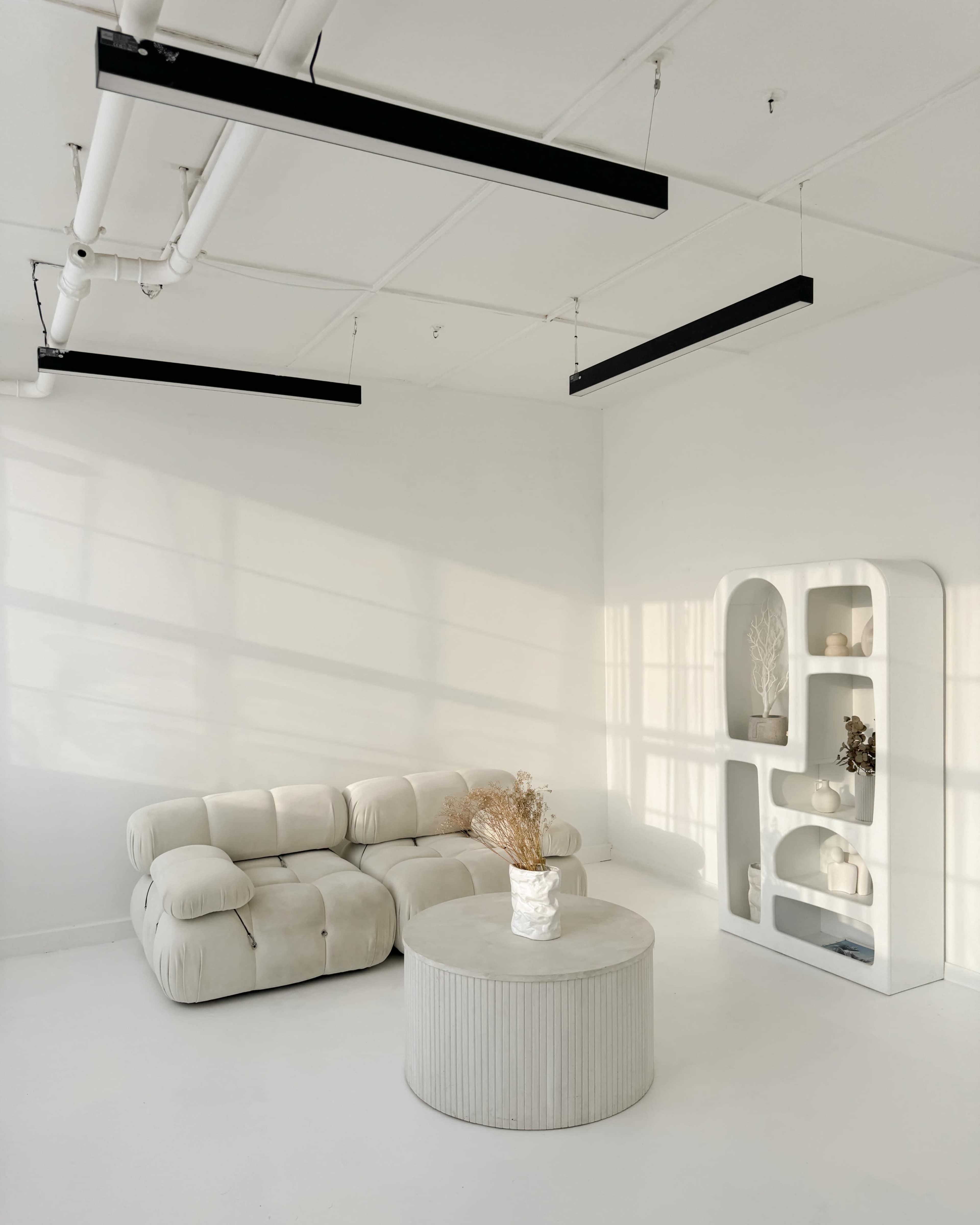 A minimalist living room features a light-colored, plush sectional sofa, a round coffee table with ribbed texture, and a white shelf displaying decorative items, all bathed in soft sunlight.