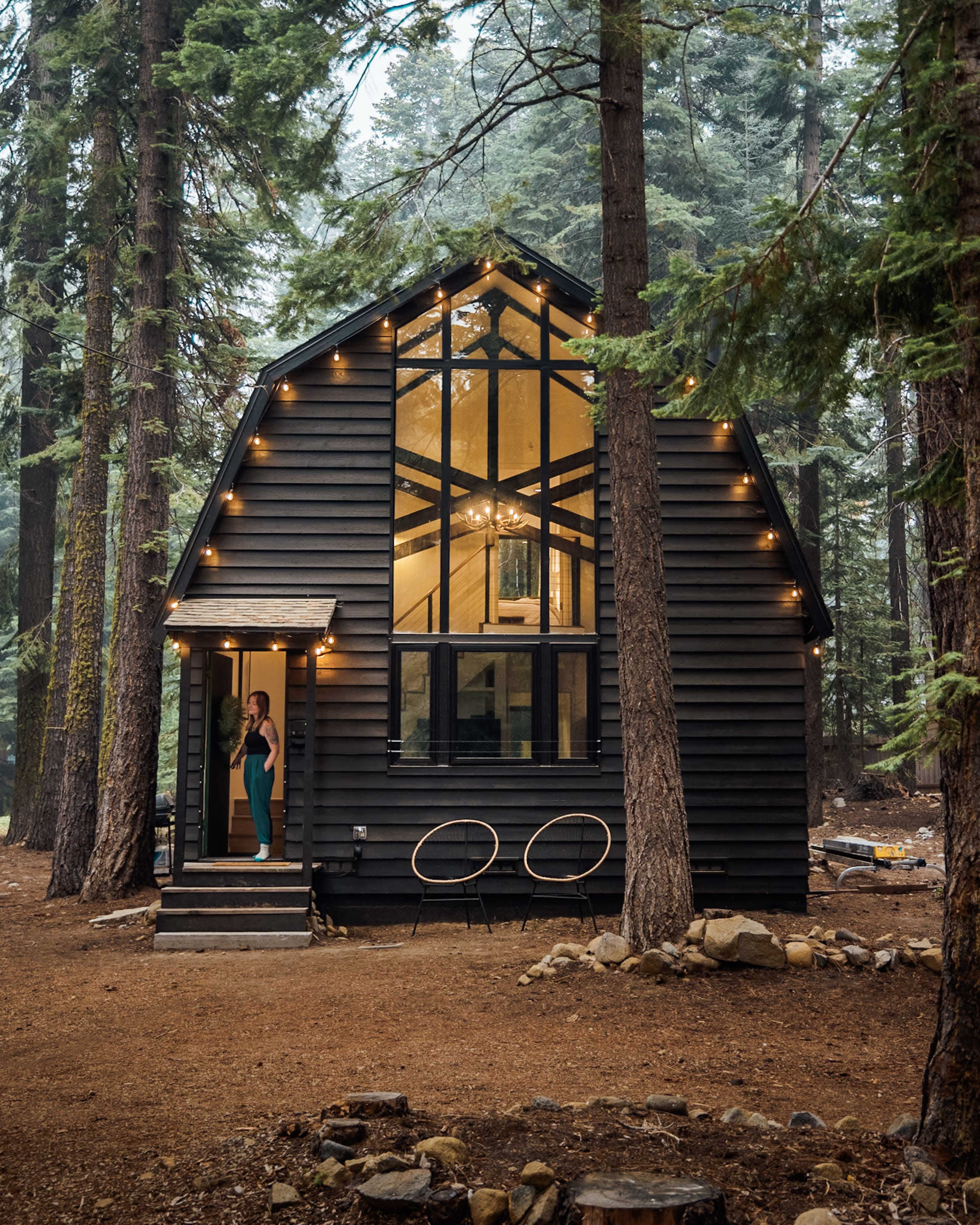 A woman stands on the porch of a modern black cabin surrounded by tall trees and a gravel pathway.