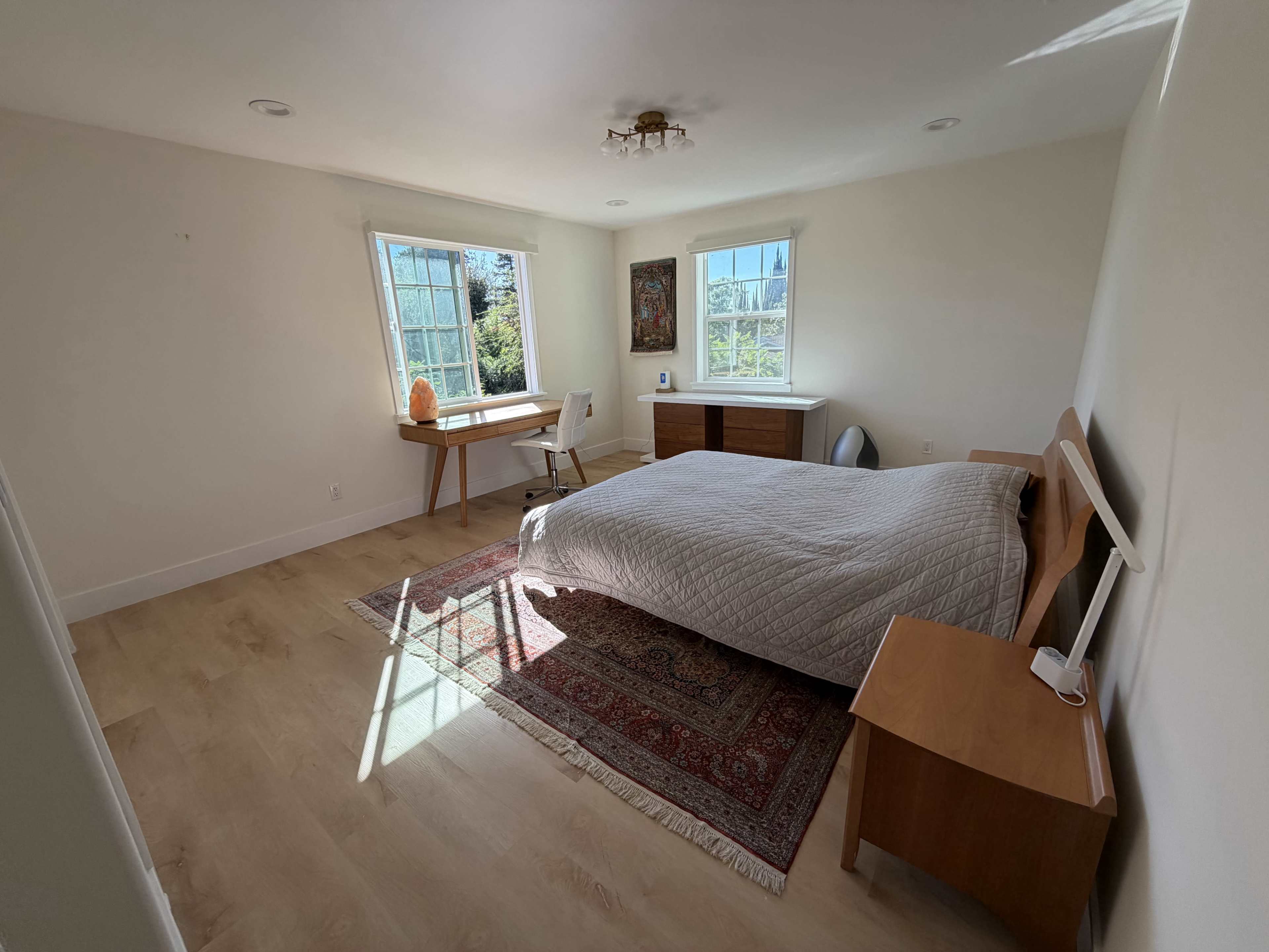 A bedroom features a bed, a desk with a chair, a dresser, and large windows allowing natural light to enter.