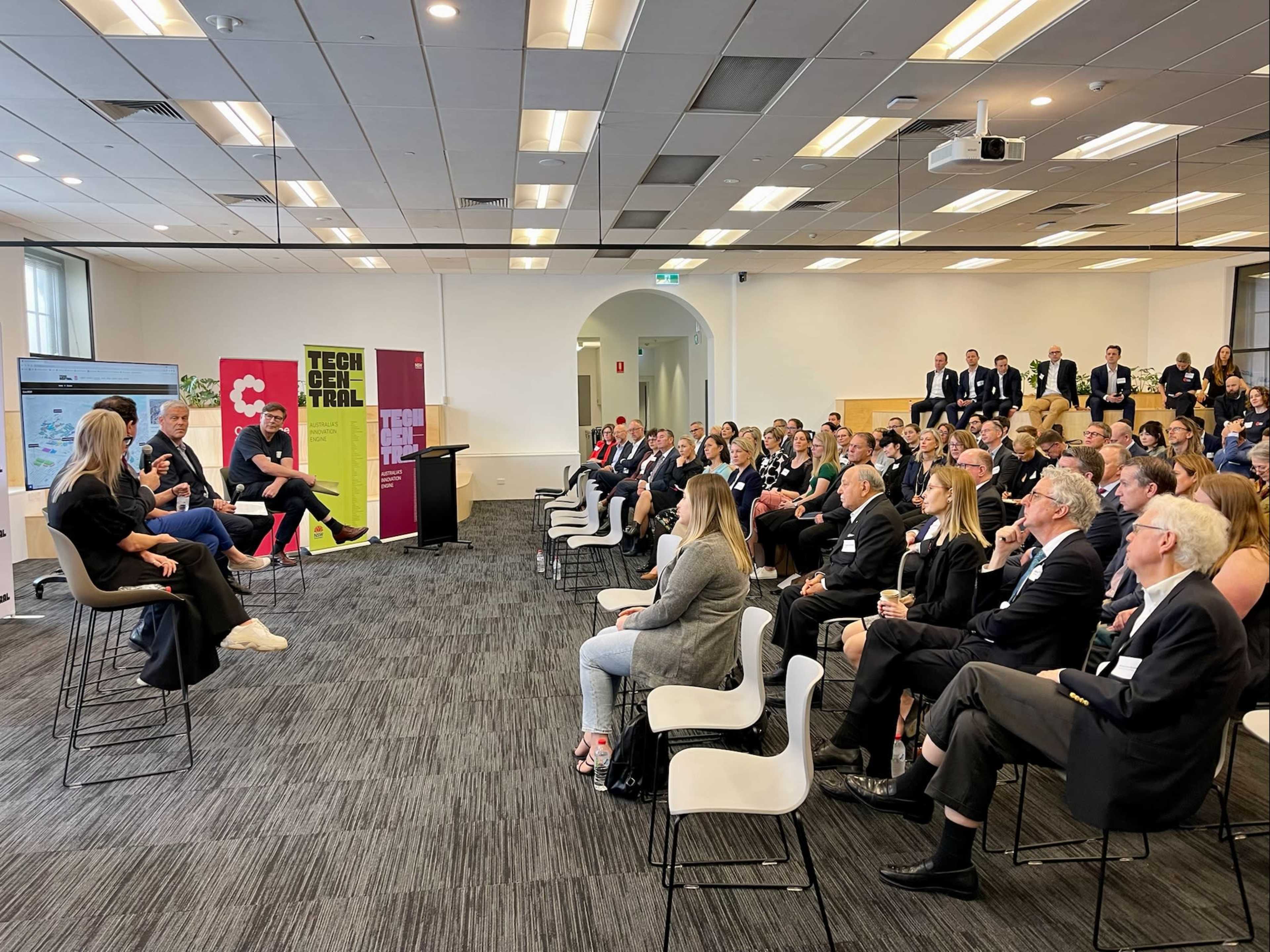 A large audience sits in a conference room while a panel discusses topics on a stage in front of them.