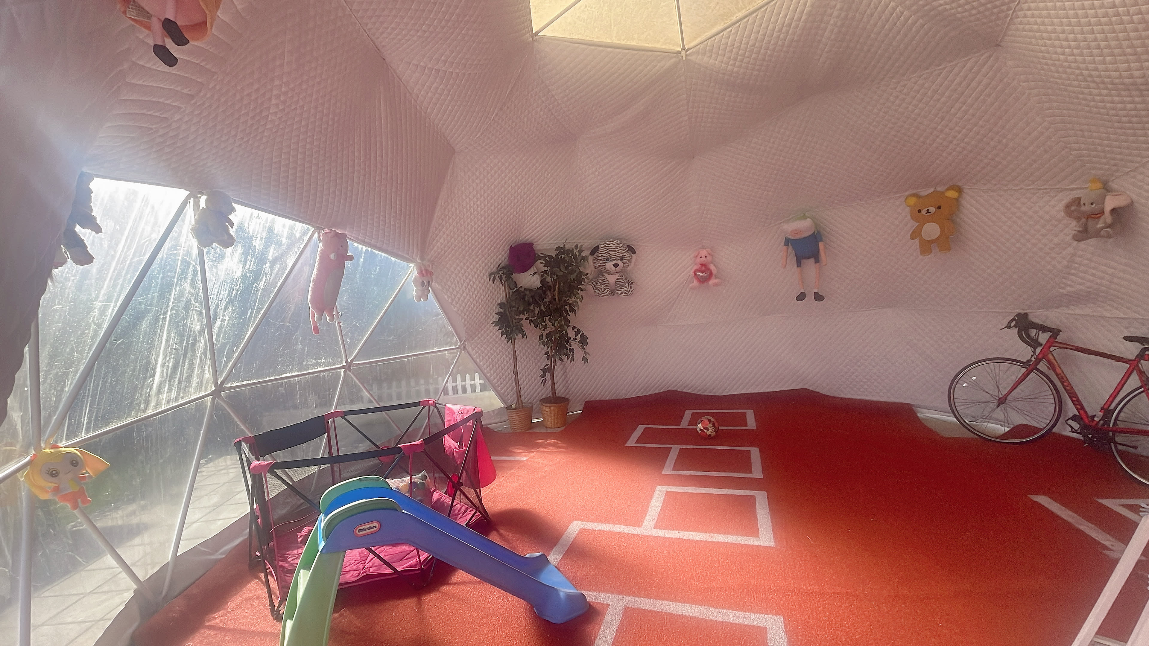 The interior of a geodesic dome playroom features a red floor with a hopscotch design, a small slide, a bike, and various plush toys hanging from the walls.