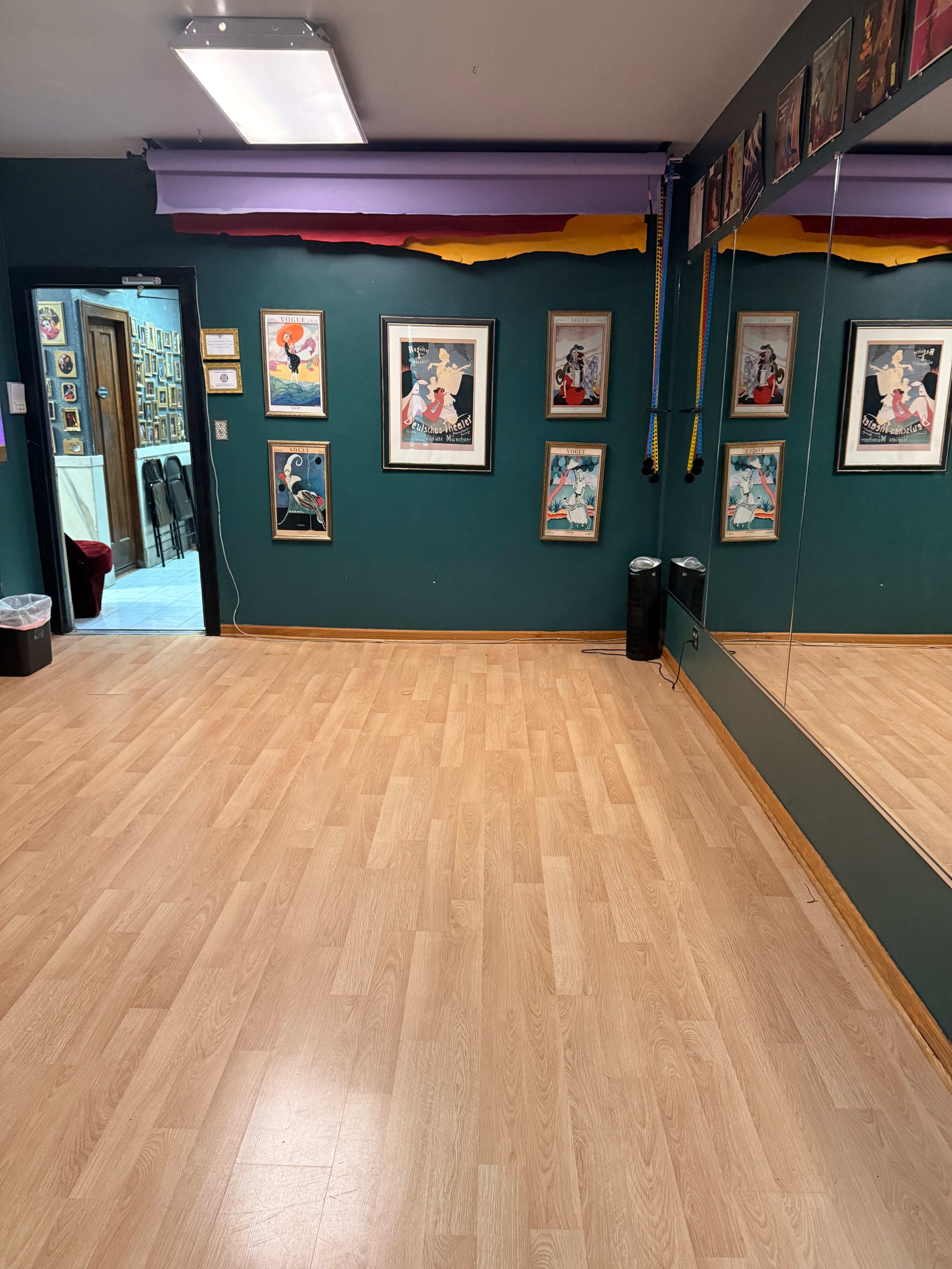 The image shows a dance studio featuring wooden flooring, green walls adorned with framed art, and a large mirror along one side.