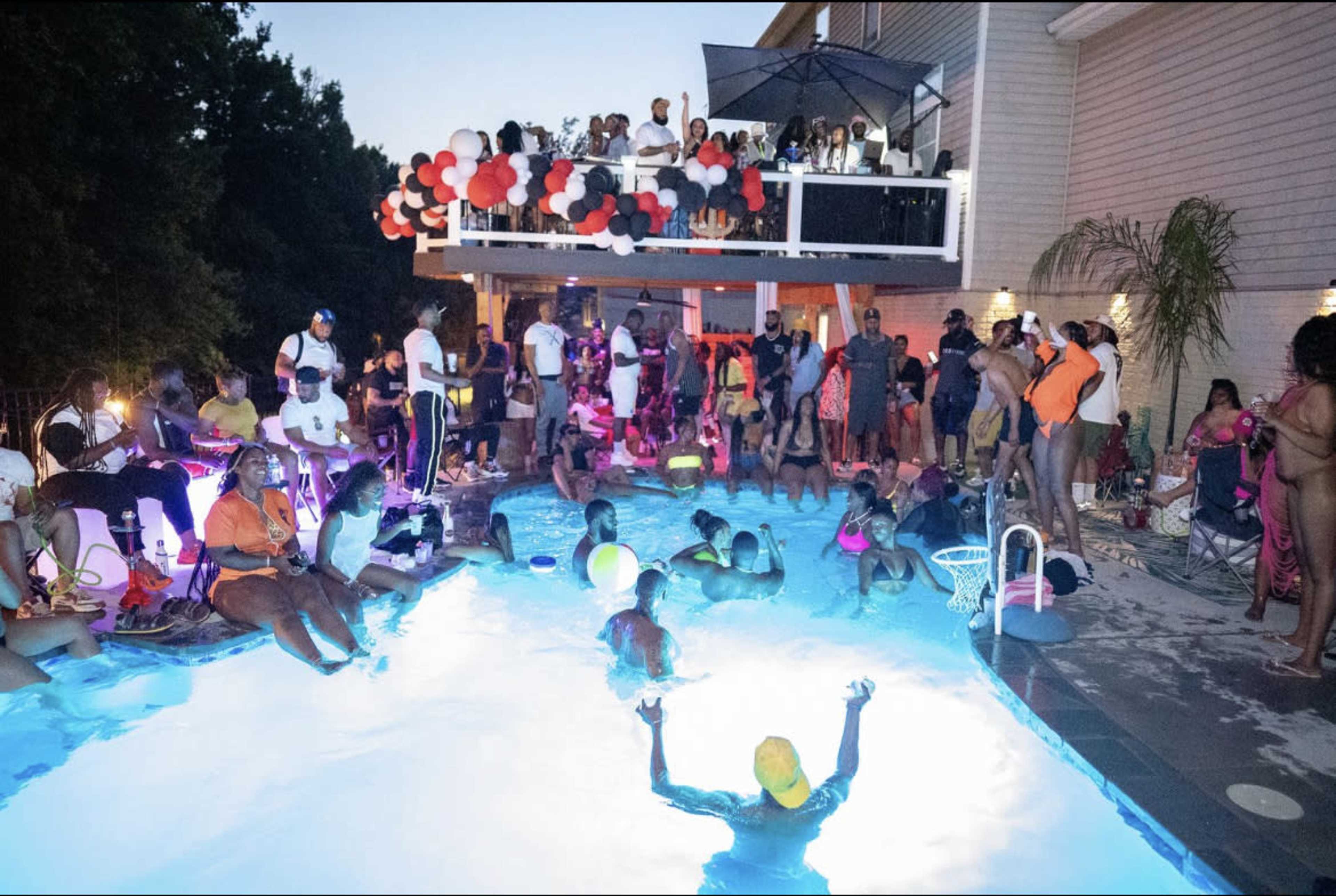 A large group of people gathers around a pool at twilight, with some in the water and others on the deck, enjoying a lively party atmosphere.