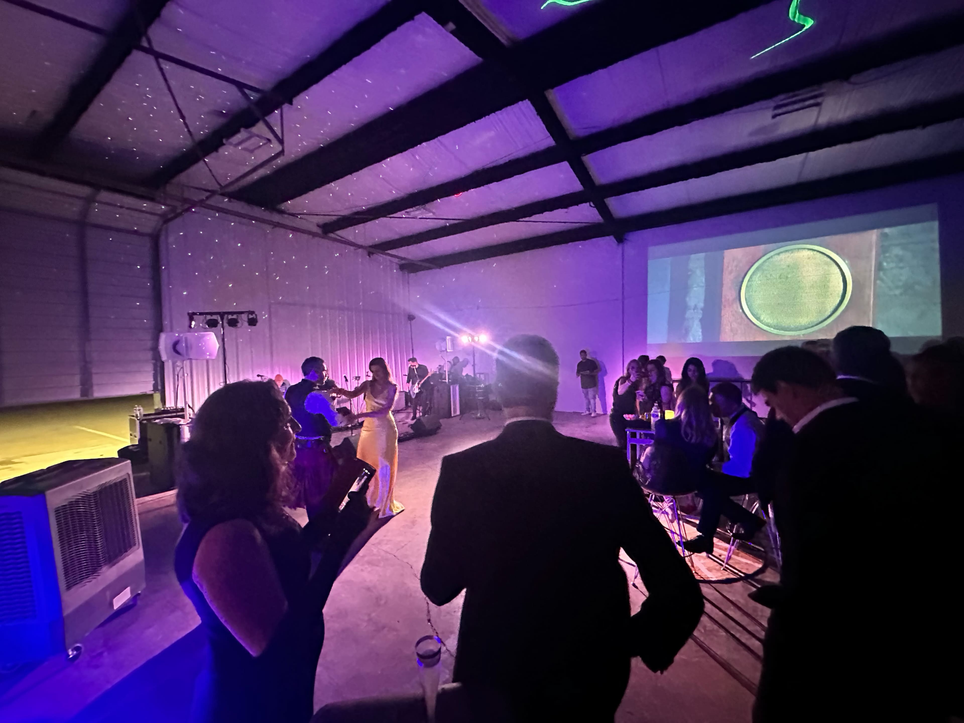 The image shows a lively indoor event in a spacious venue, with guests mingling and an entertainer performing on stage, illuminated by colorful lights.