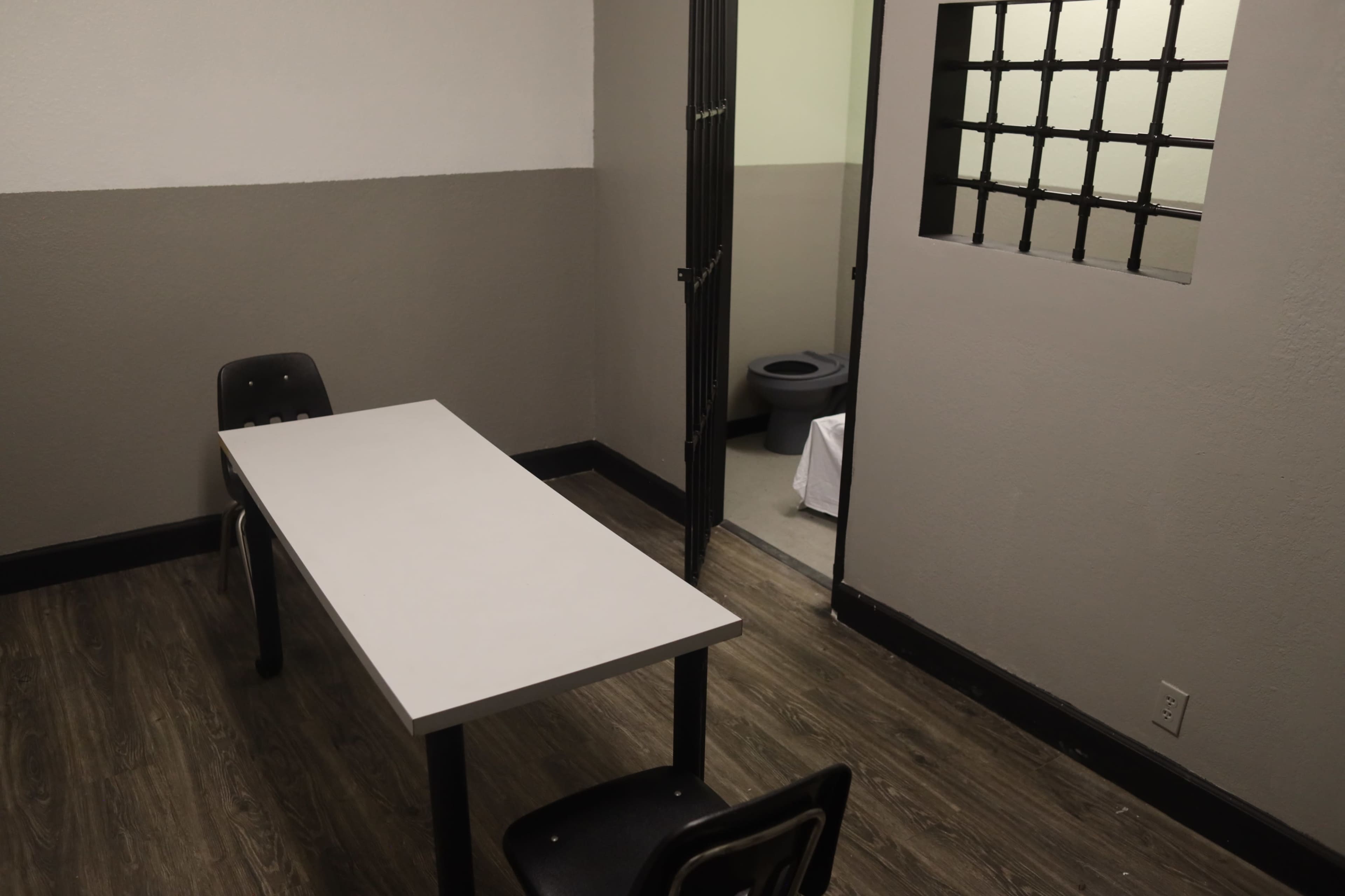 The image shows a small, minimally furnished room with a table and two chairs, featuring a barred window and an adjacent area containing a toilet.