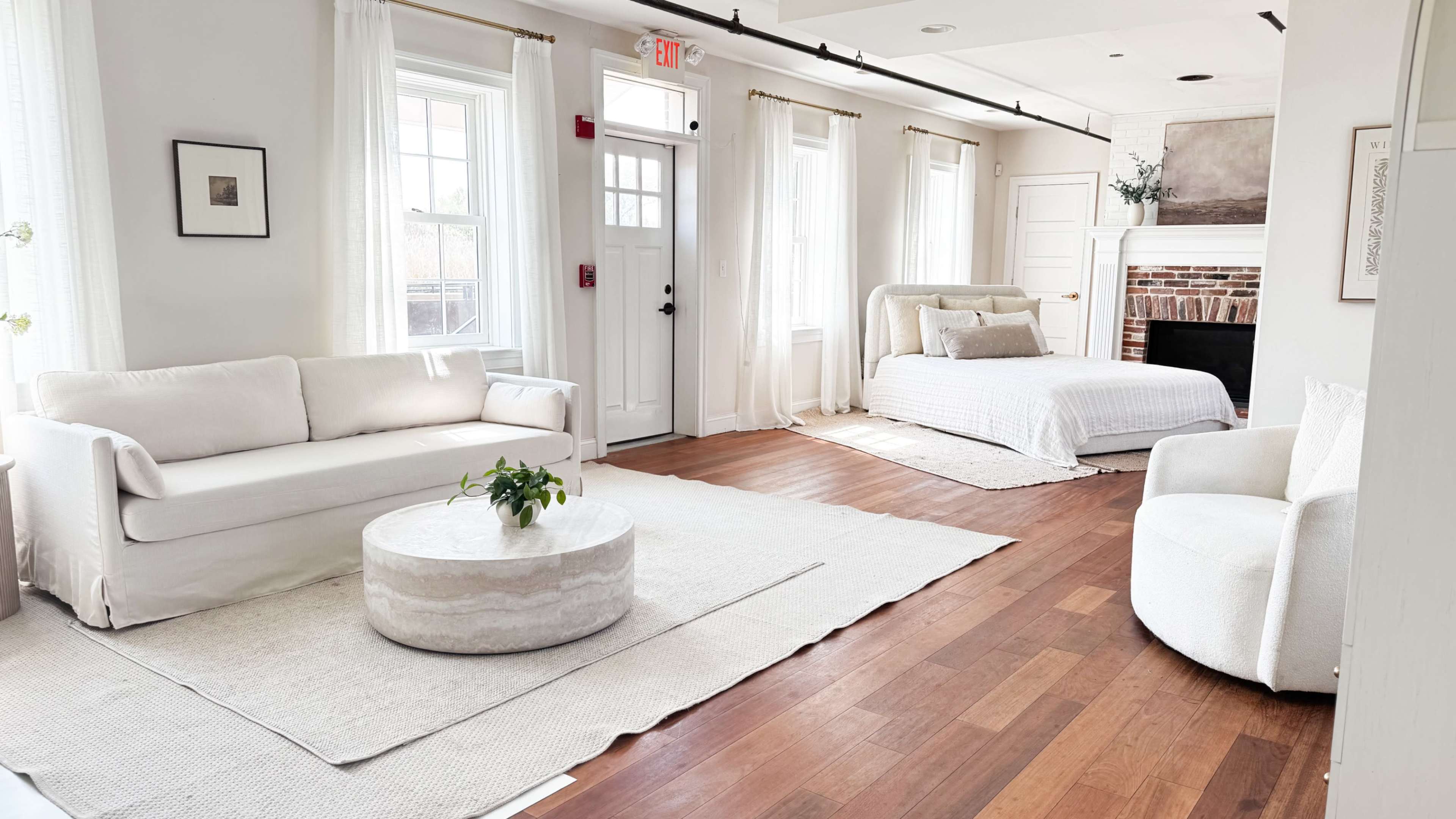 The image shows a bright, modern living space featuring a light-colored sofa, a round coffee table, and a bed with a simple bedspread against a backdrop of large windows and hardwood floors.