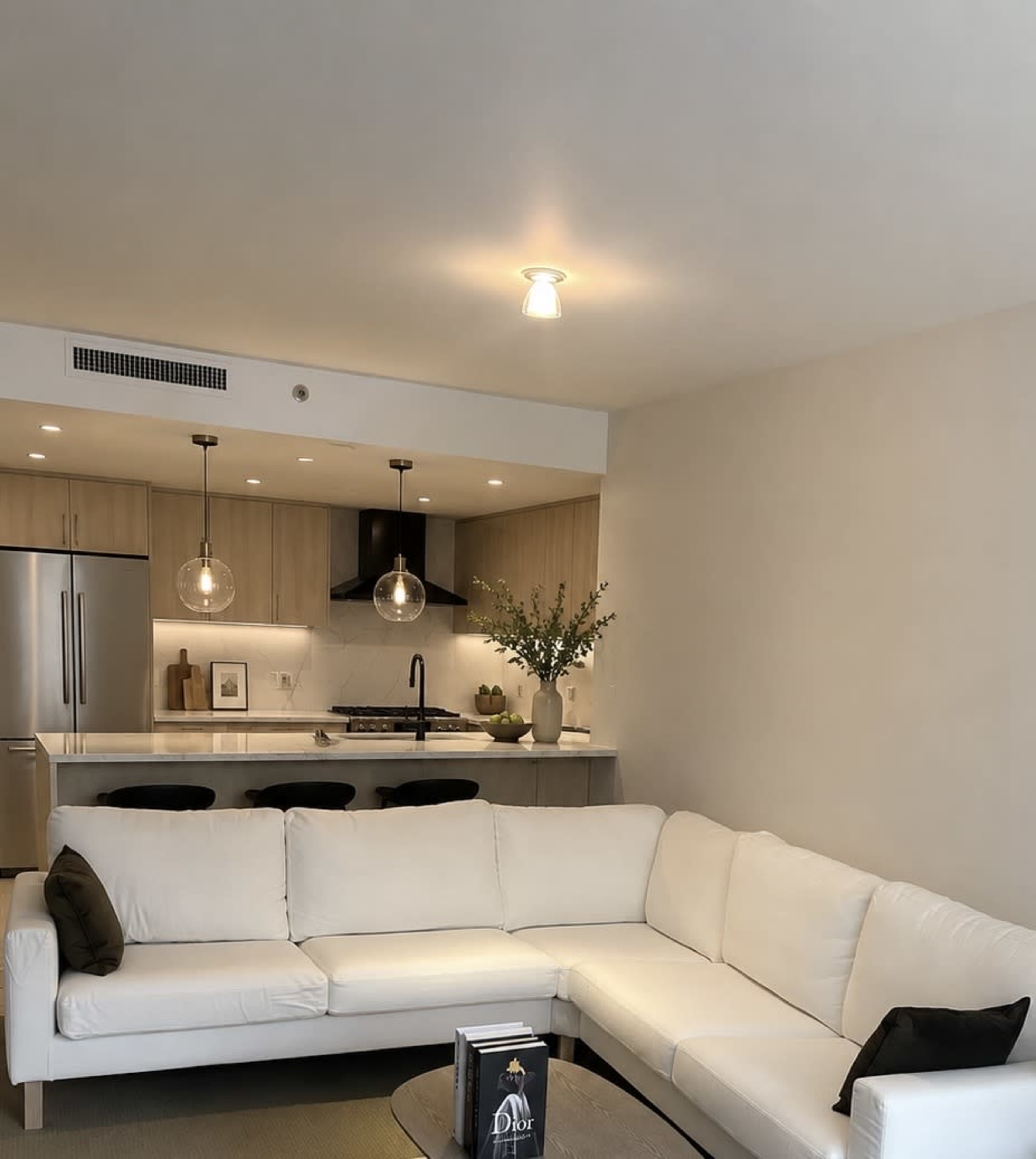The image shows a modern living room with a white sectional sofa, a round coffee table, and a kitchen area featuring wooden cabinetry and pendant lighting.