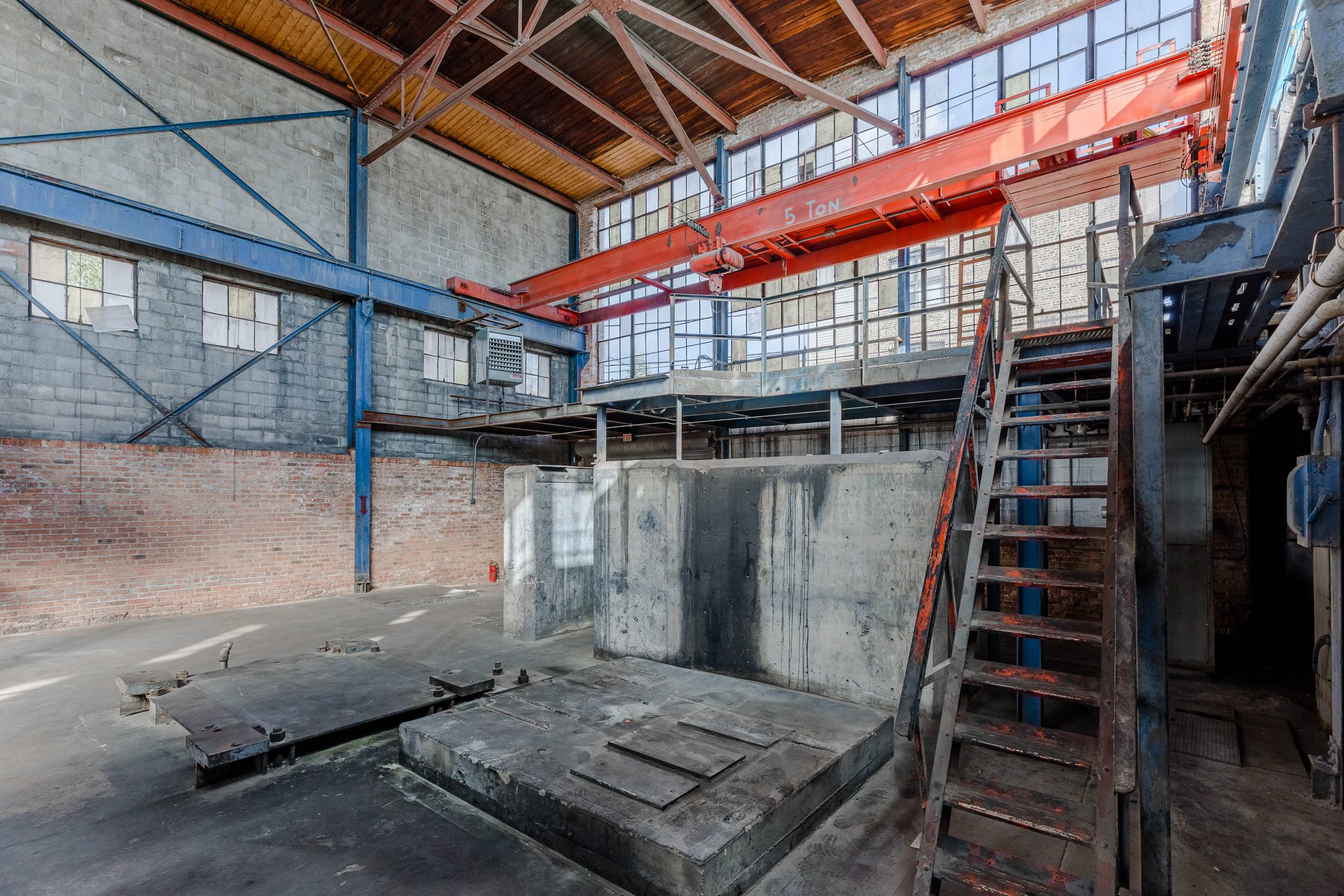 An industrial interior with a large overhead crane, concrete structures, and a metal staircase against brick walls and large windows.