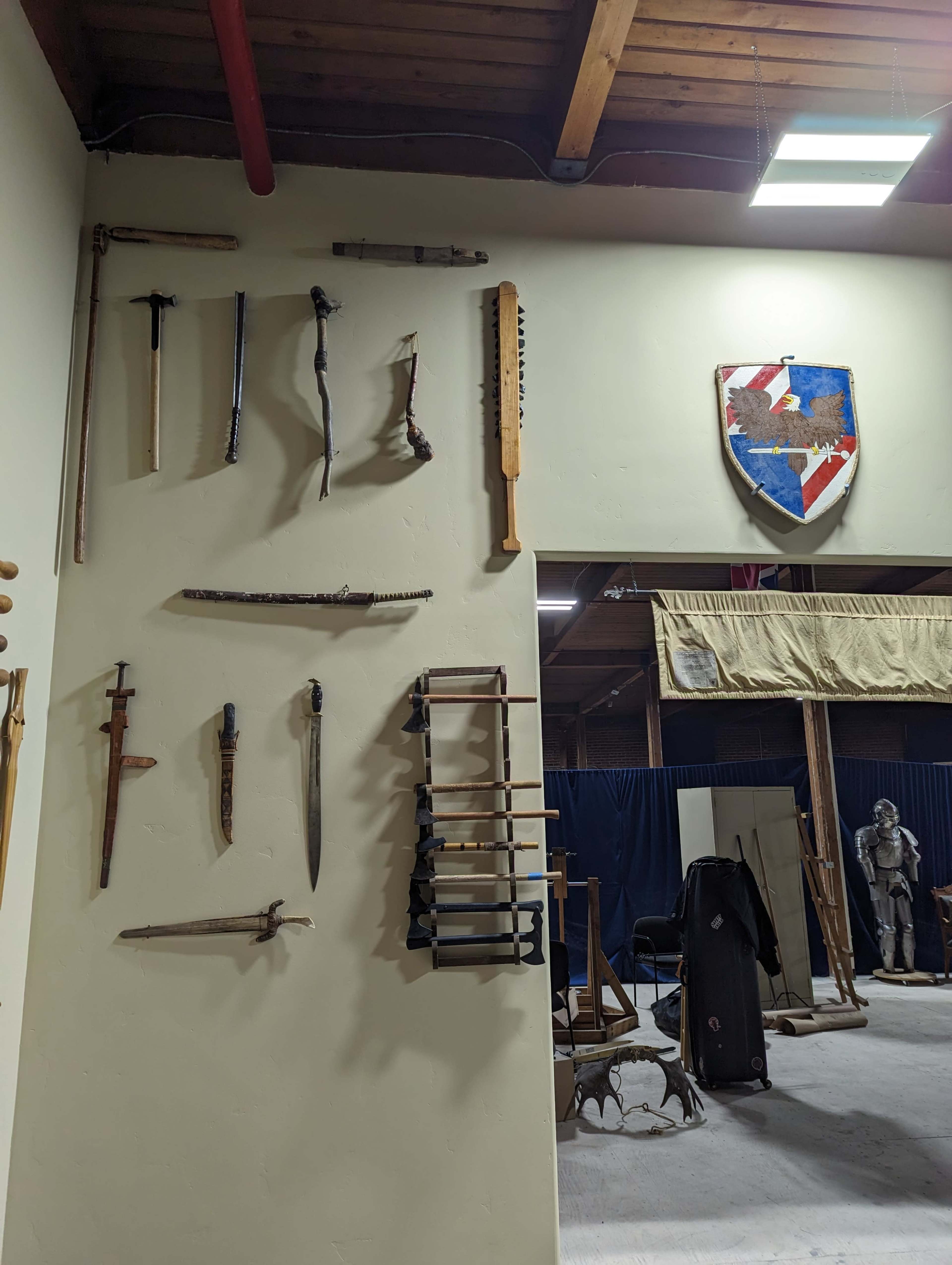 A wall displaying various historical weapons and a shield, with additional artifacts and a draped area visible in the background.