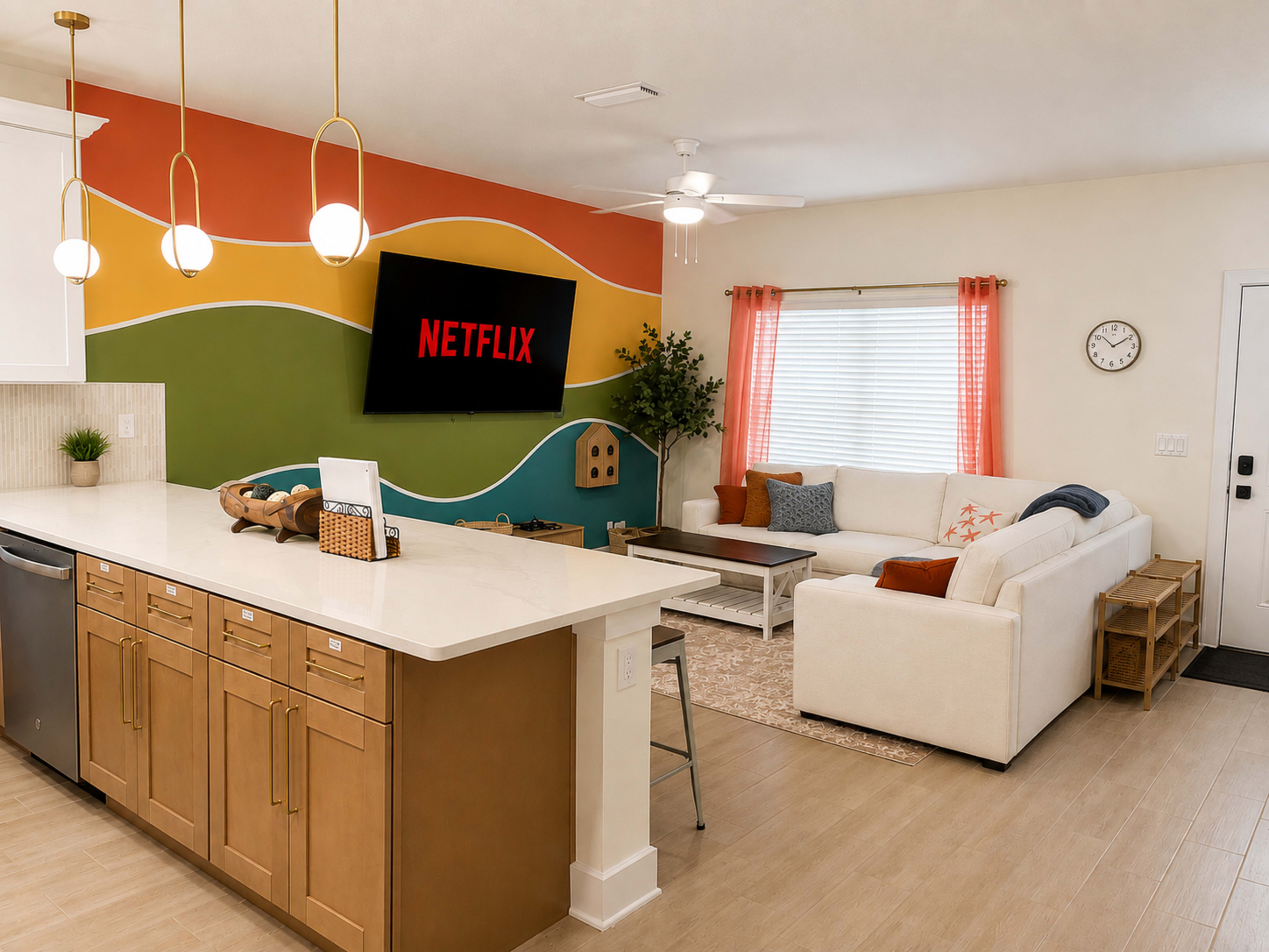 A modern living area features a white sectional couch, a colorful accent wall with a Netflix display, and a kitchen island with wooden cabinetry.