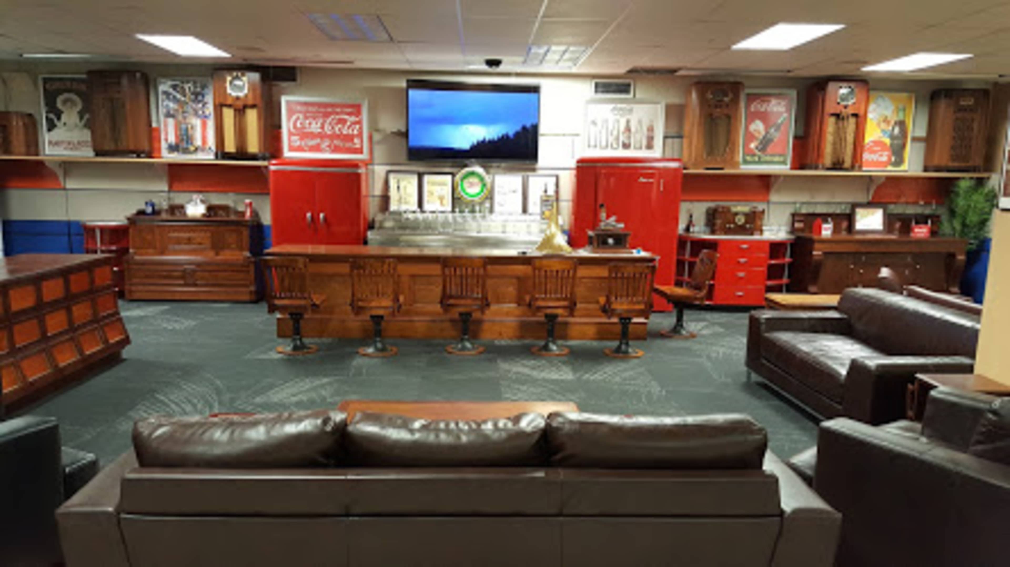 A retro-style lounge area featuring a bar with seating, vintage Coca-Cola memorabilia, a large television, and wooden cabinets in a spacious room.