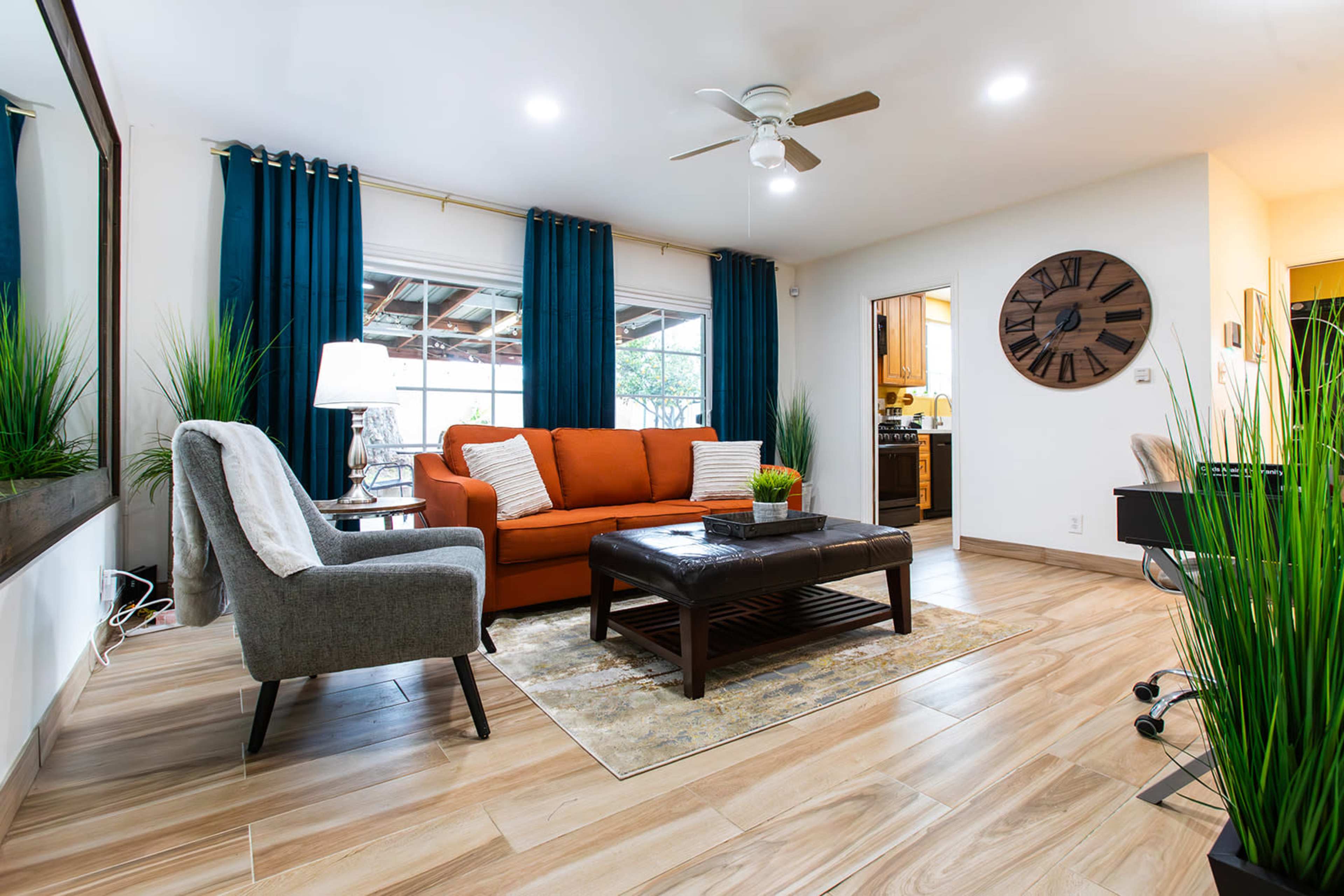 A living room featuring an orange sofa, a gray armchair, a coffee table on a patterned rug, and potted plants, with a kitchen visible in the background.
