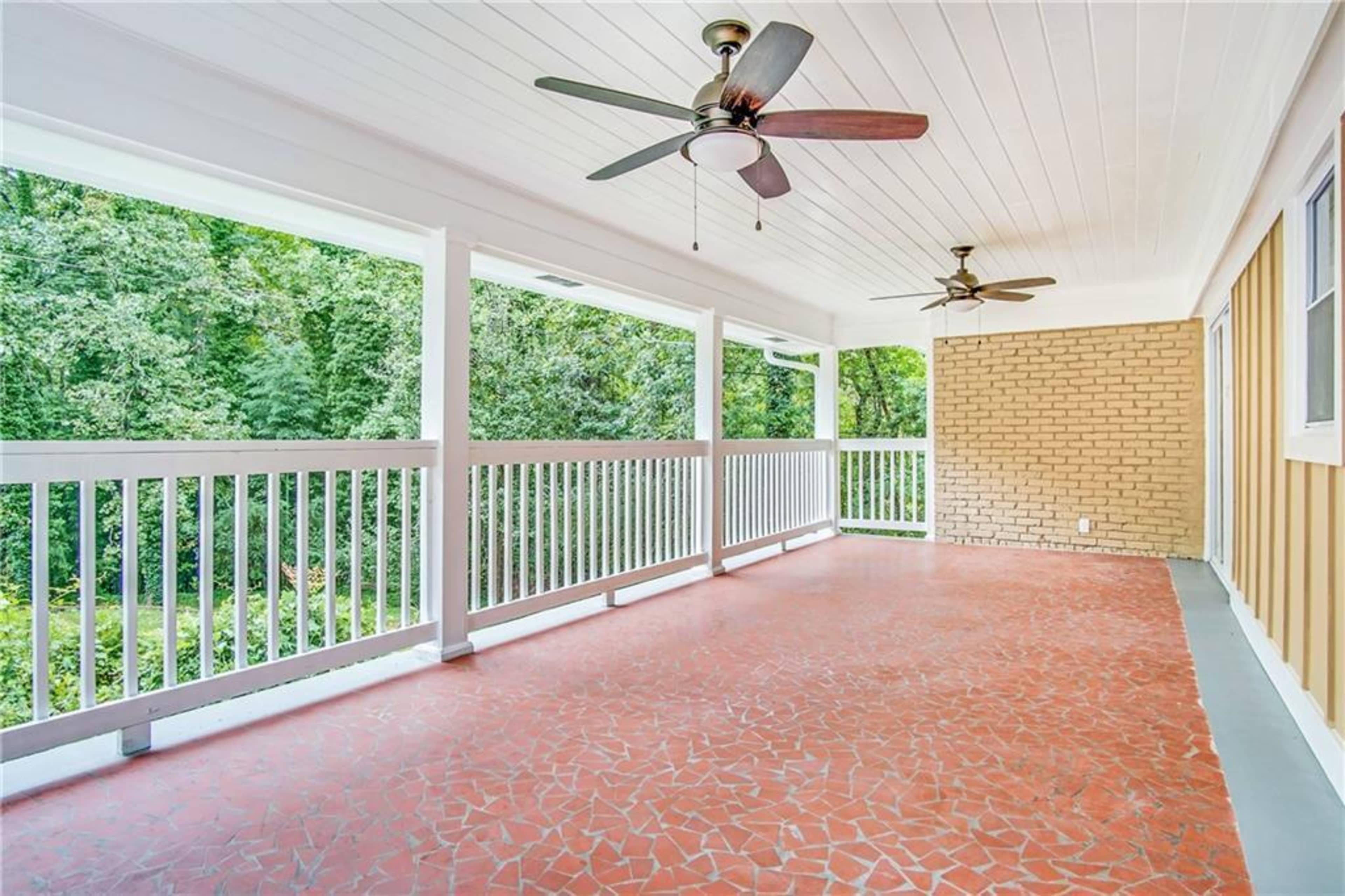 A spacious covered porch with a brick wall, two ceiling fans, and a view of greenery beyond the railing.