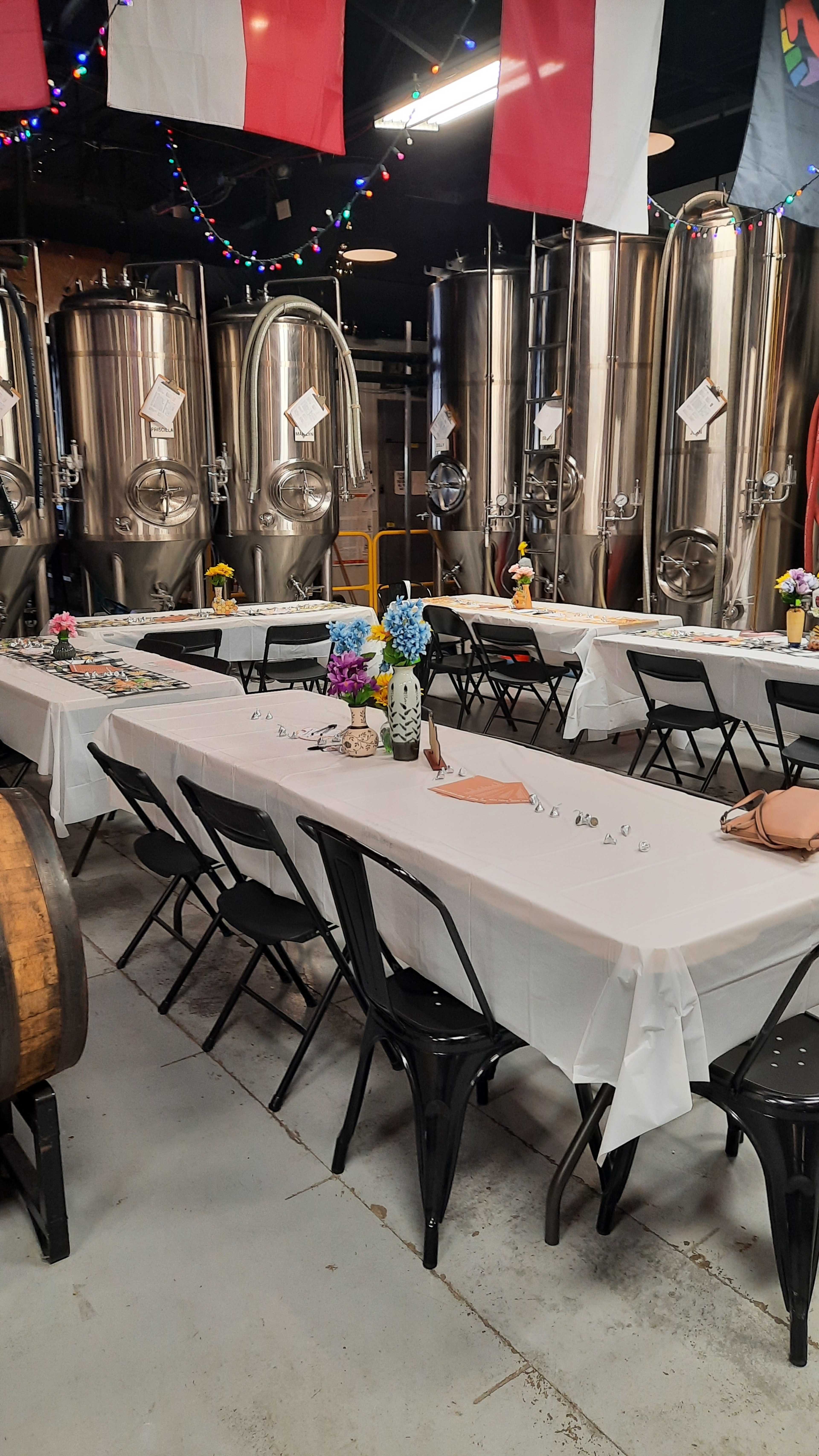 A long, white-tableclothed dining table is set up in a brewery, surrounded by large stainless steel fermentation tanks and decorated with flowers.