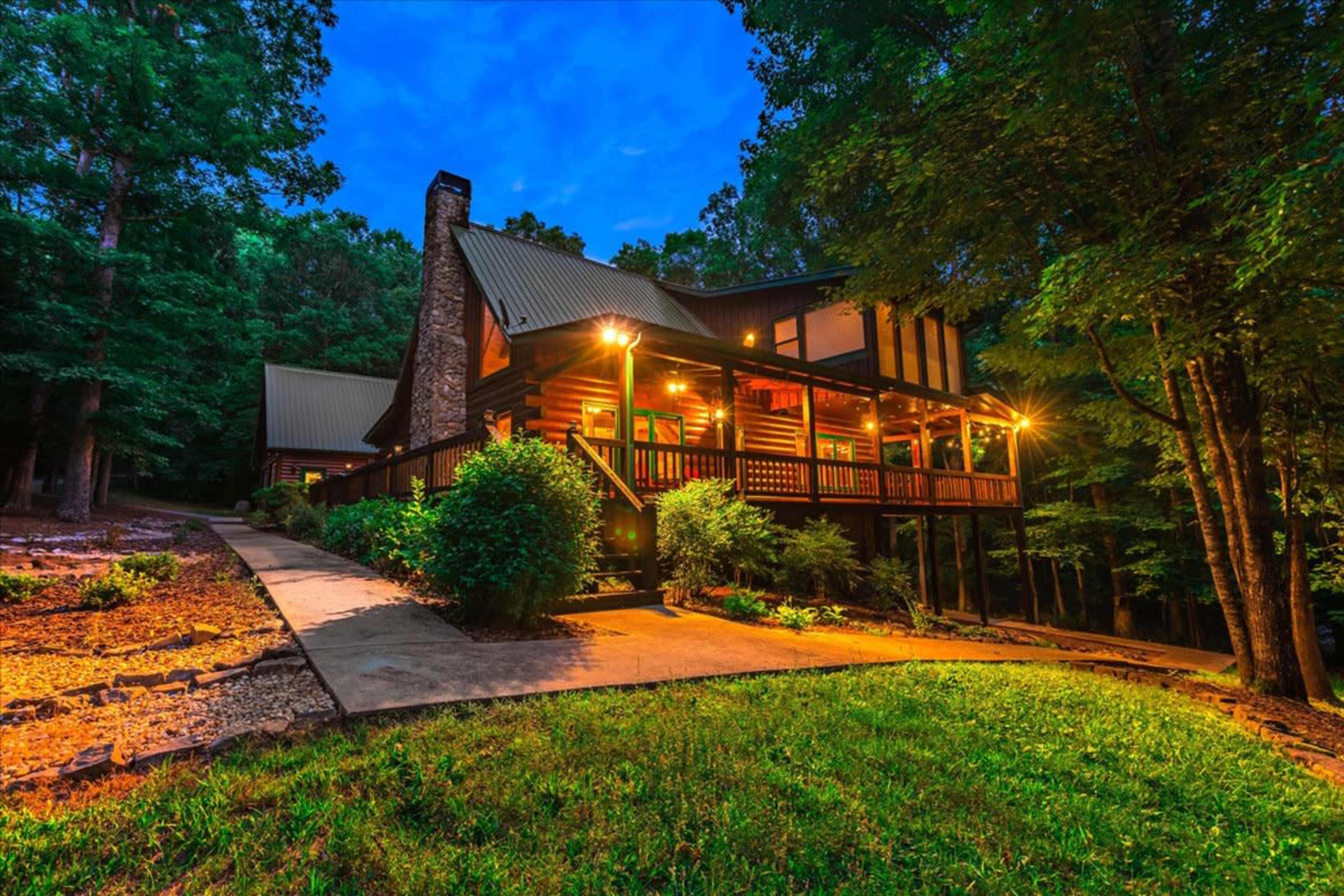 A large log cabin with warm lighting is situated among trees on a winding driveway in a wooded area.