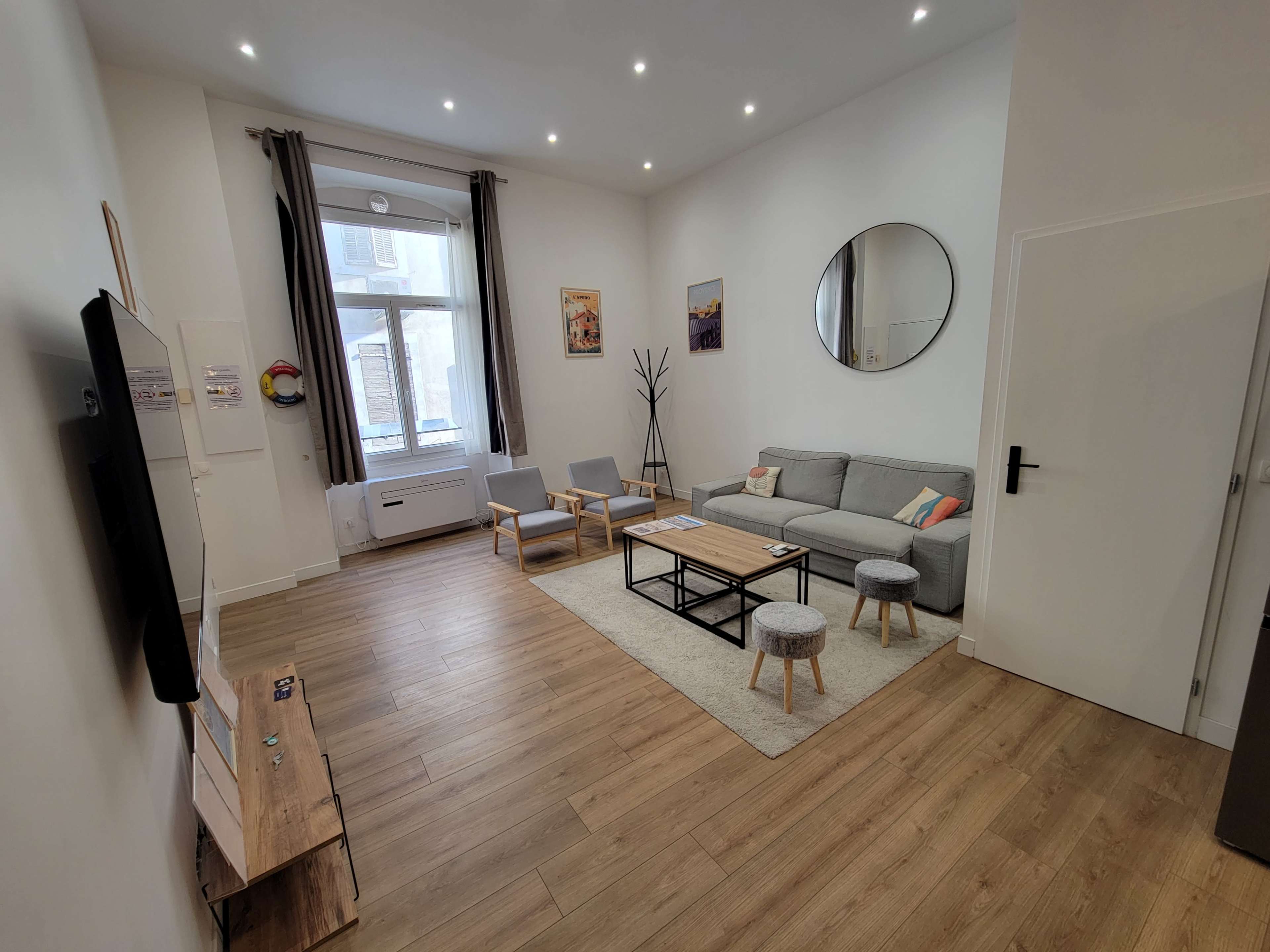 The image shows a modern, well-lit living room featuring a gray sofa, two armchairs, a coffee table, and a wall-mounted TV.