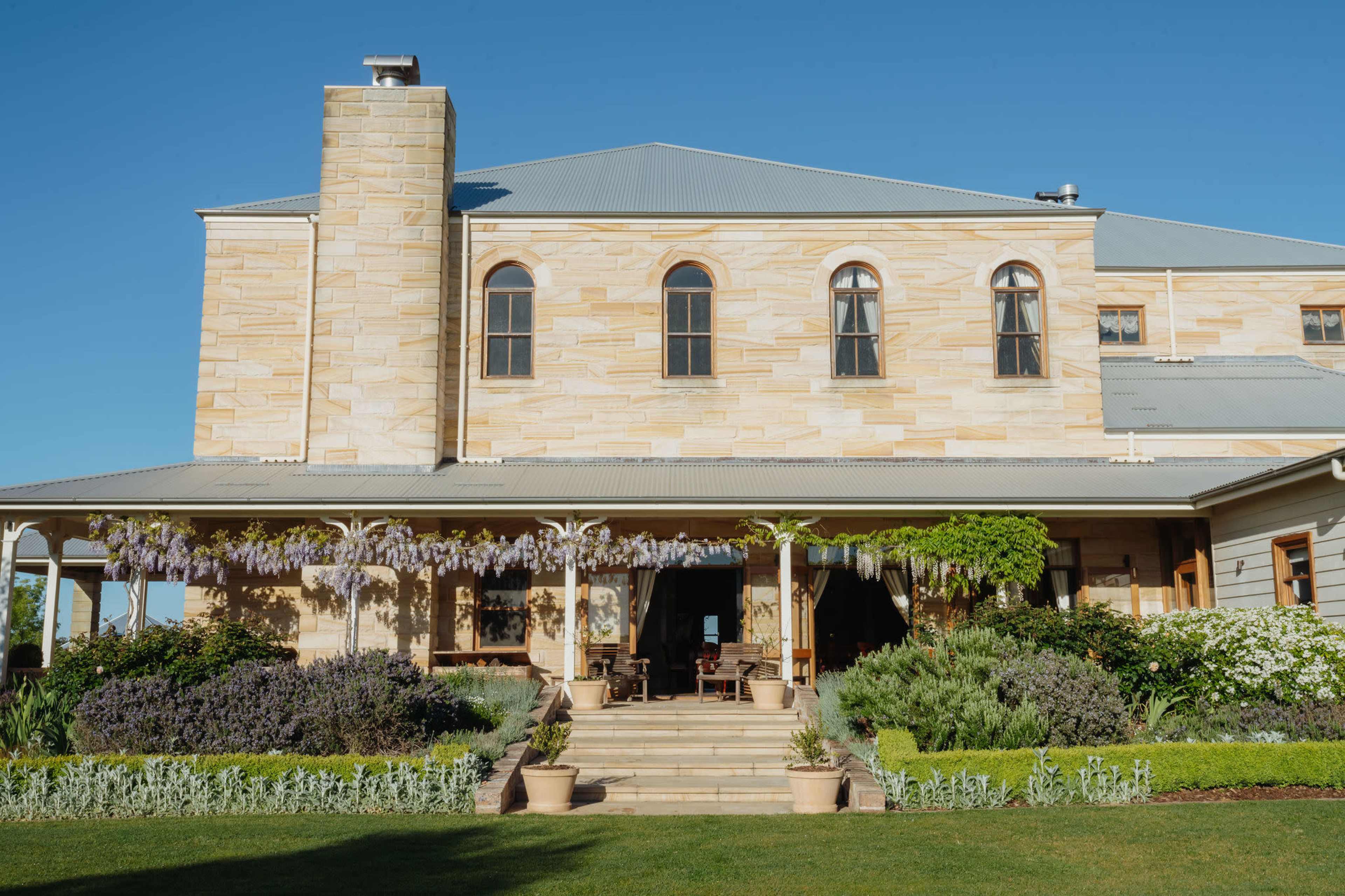 The image depicts a large stone house with multiple windows, featuring a covered porch adorned with flowering plants and a well-manicured garden.