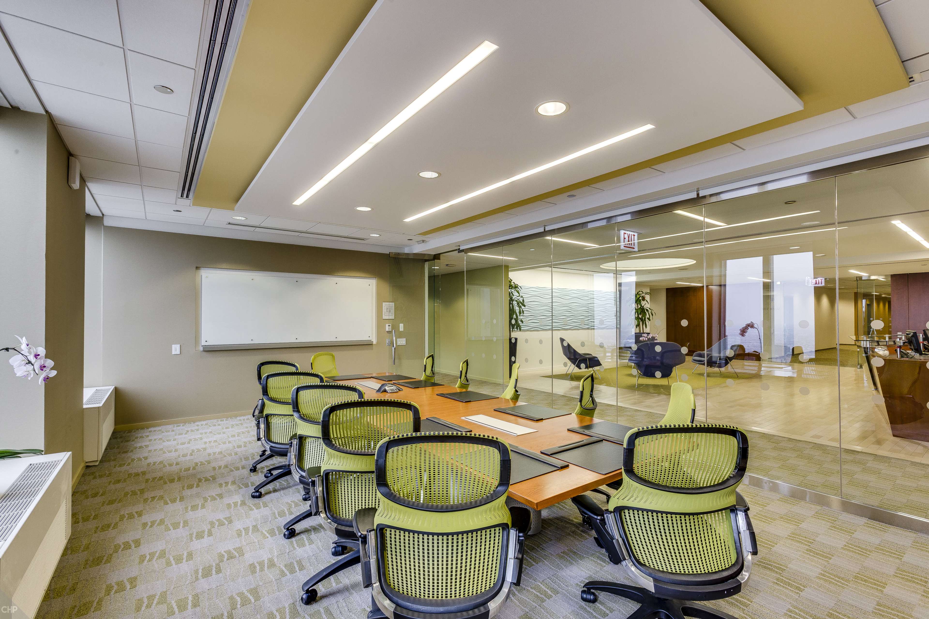 A modern conference room features a long wooden table surrounded by green ergonomic chairs, with a whiteboard and glass walls.