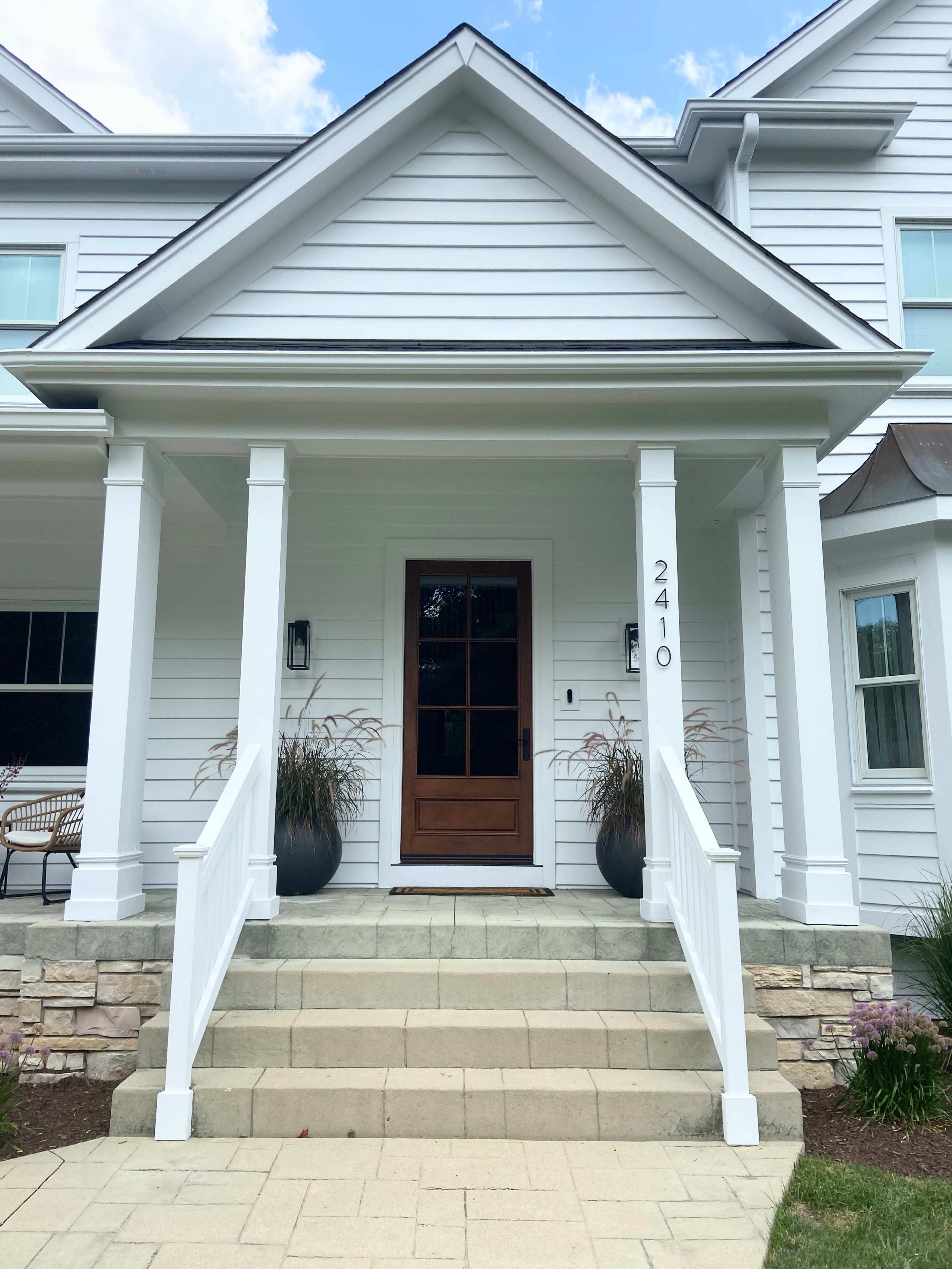 The image shows the front entrance of a white house with a wooden door and stone steps, flanked by two large planters.