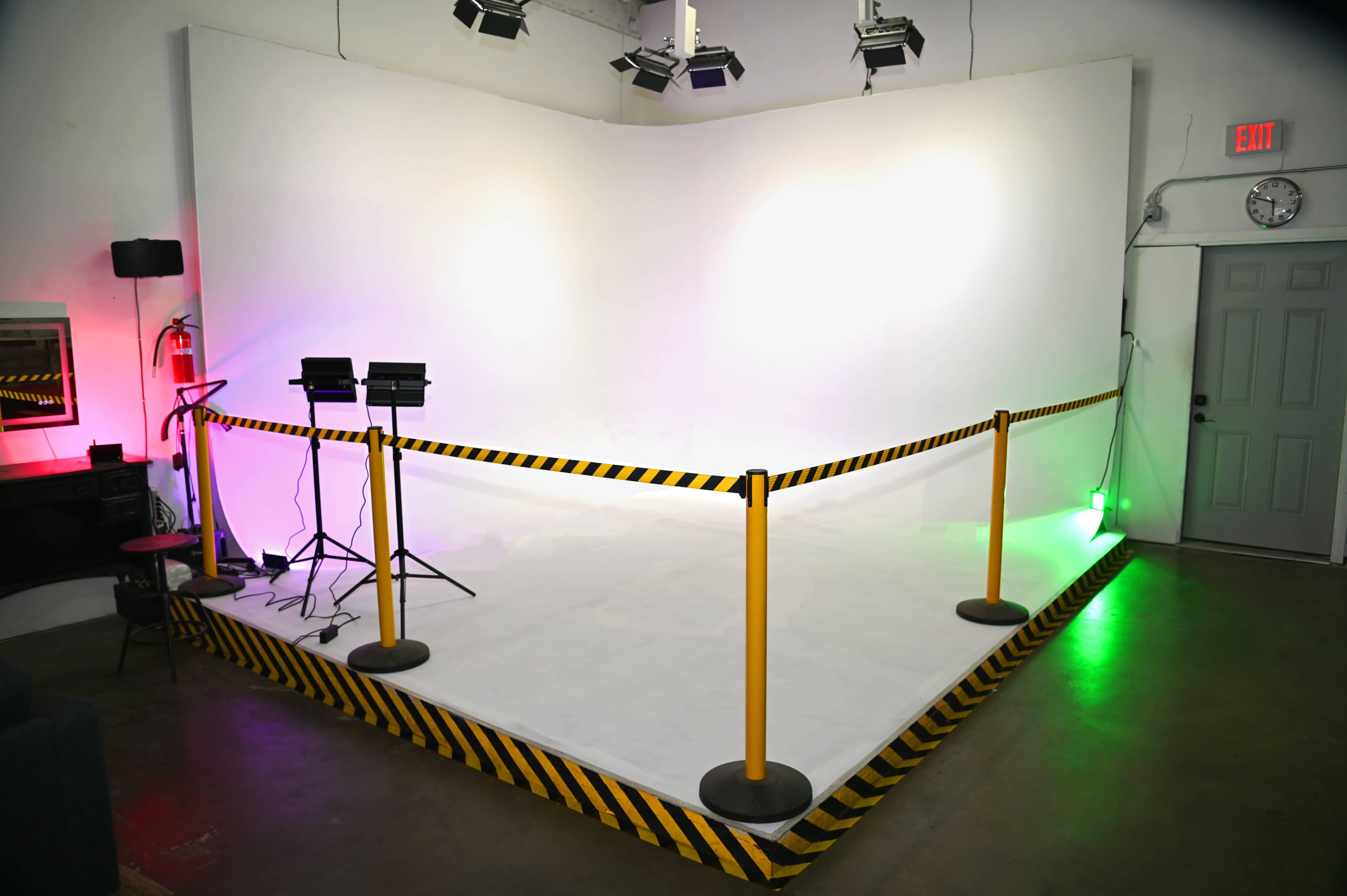 The image shows a studio space with a white backdrop, bordered by yellow and black safety barriers, and illuminated by colored lights.