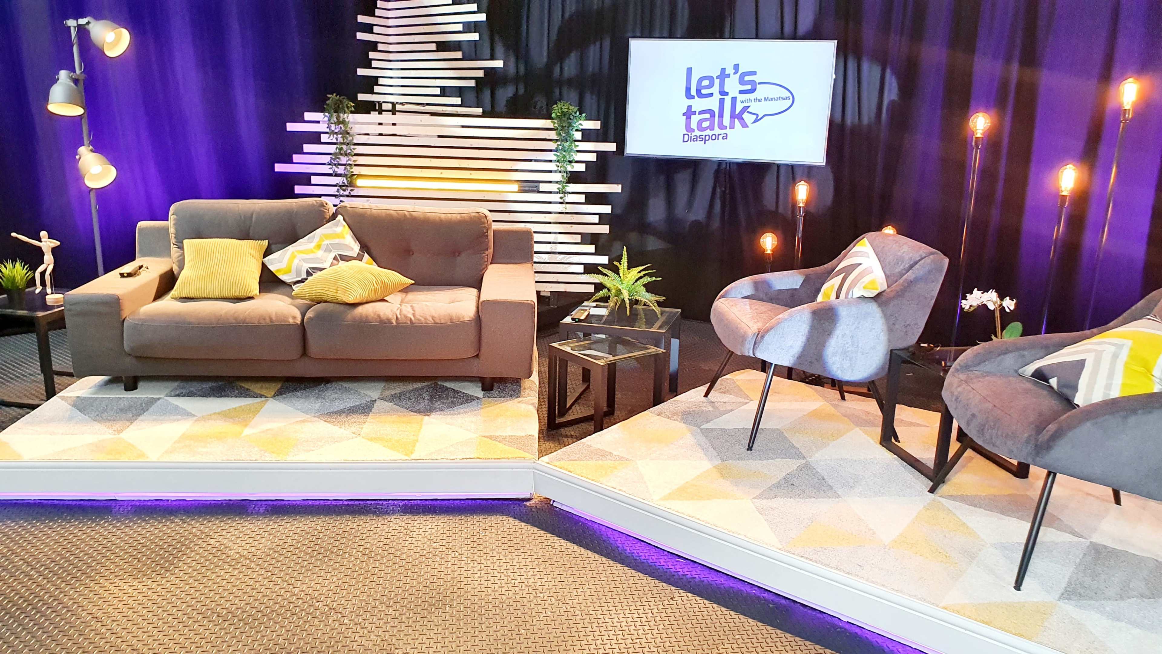 A modern talk show set featuring a gray sofa, two accent chairs, decorative plants, and a backdrop with purple lighting and a screen displaying "Let's Talk Diaspora."