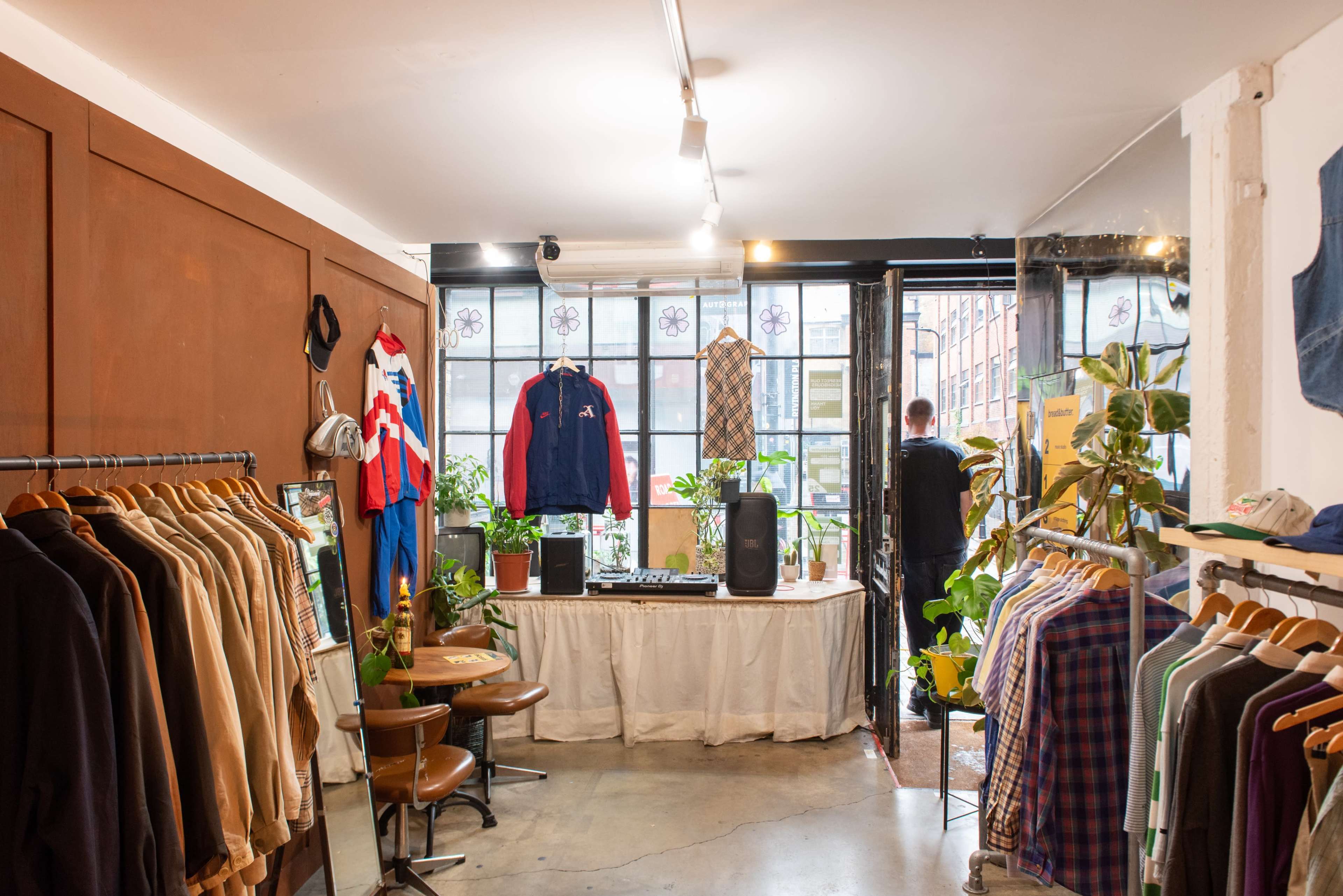 The image shows the interior of a clothing store with racks of apparel, a DJ setup, and a large window that opens to the outside.