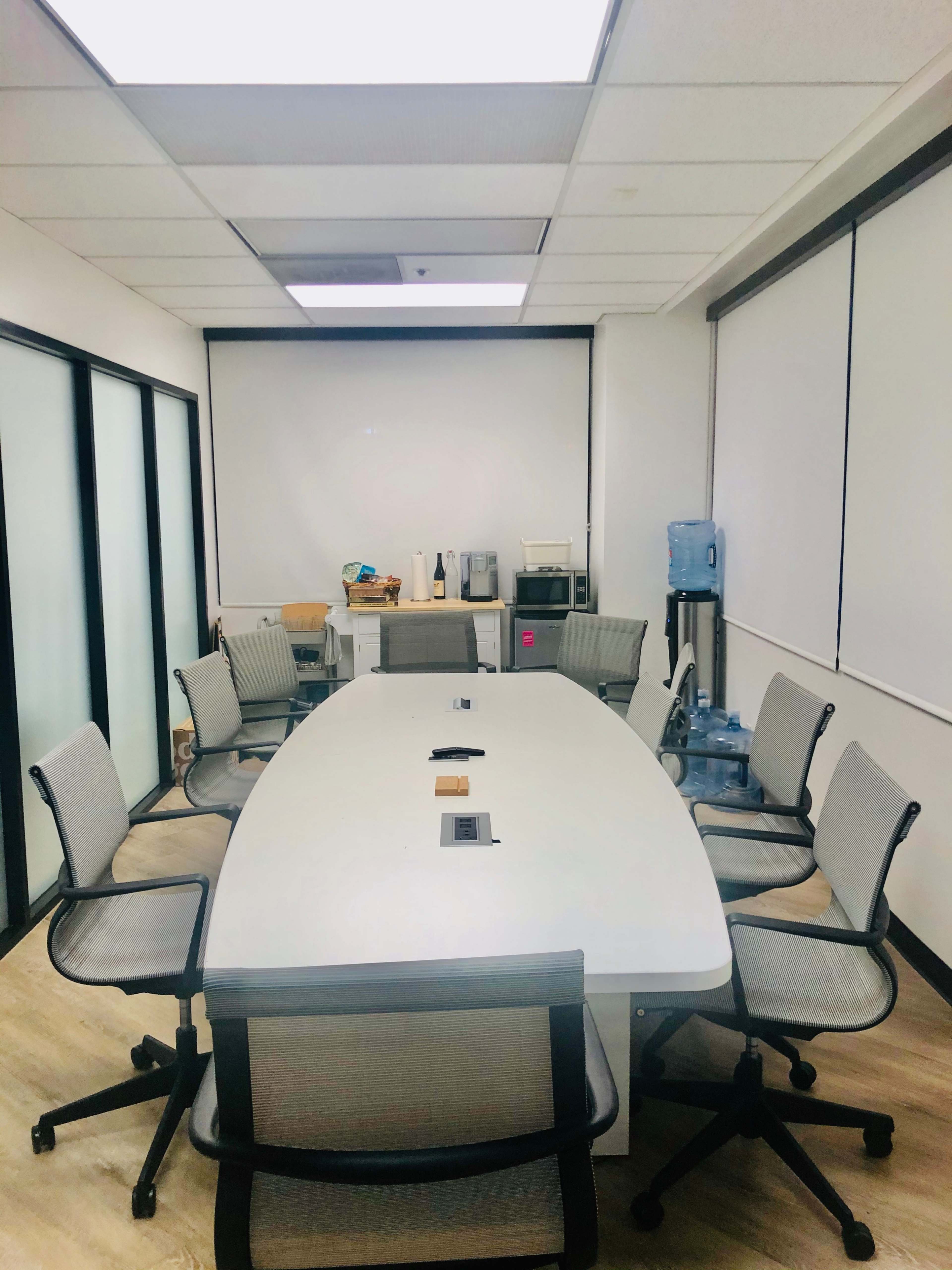 A conference room features a large oval table surrounded by six chairs, with a kitchenette and a water cooler in the background.