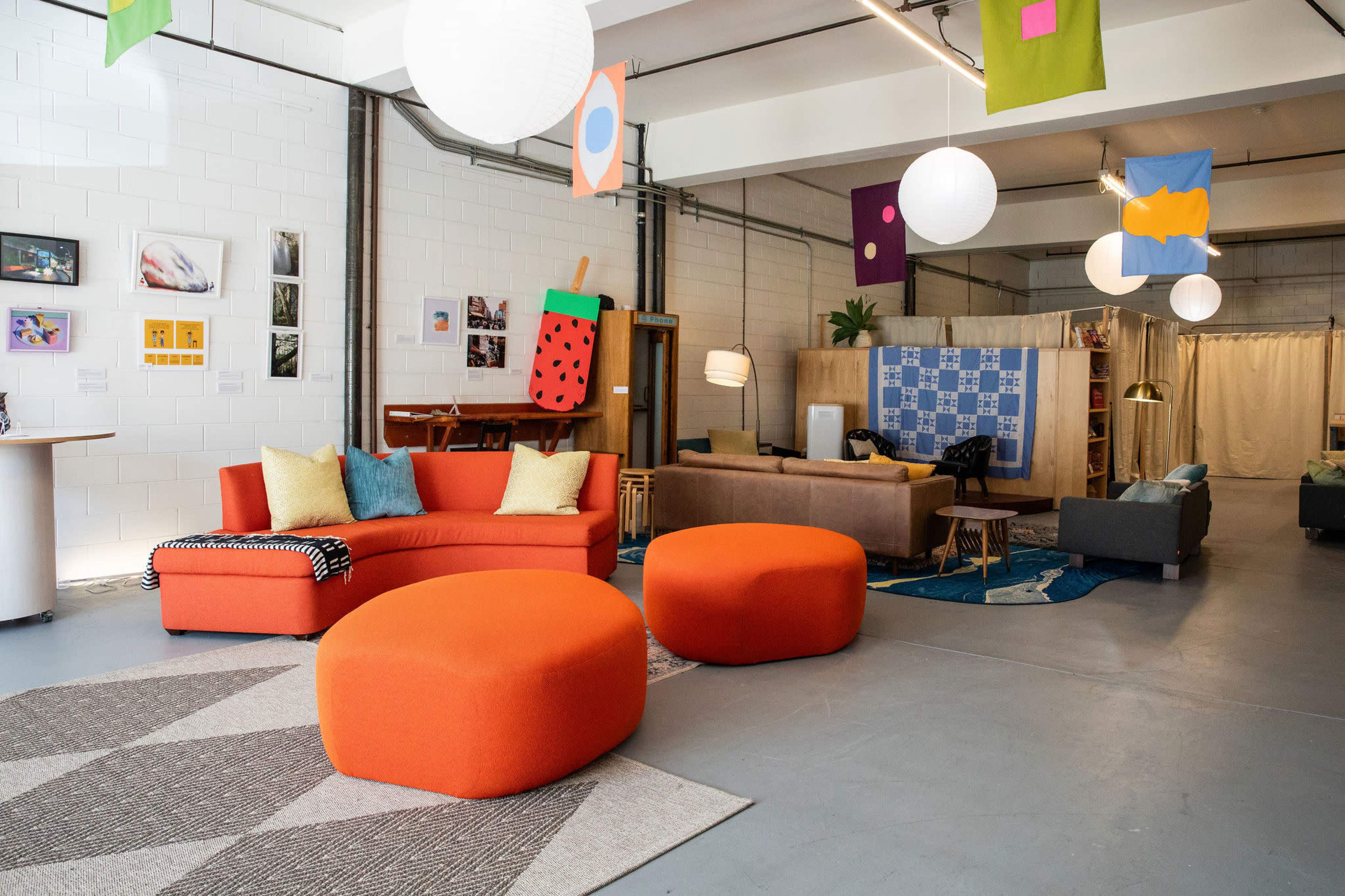 The image shows a modern, open space featuring colorful furniture, including an orange sectional sofa and round ottomans, along with various artworks and decorations on the walls and hanging from the ceiling.