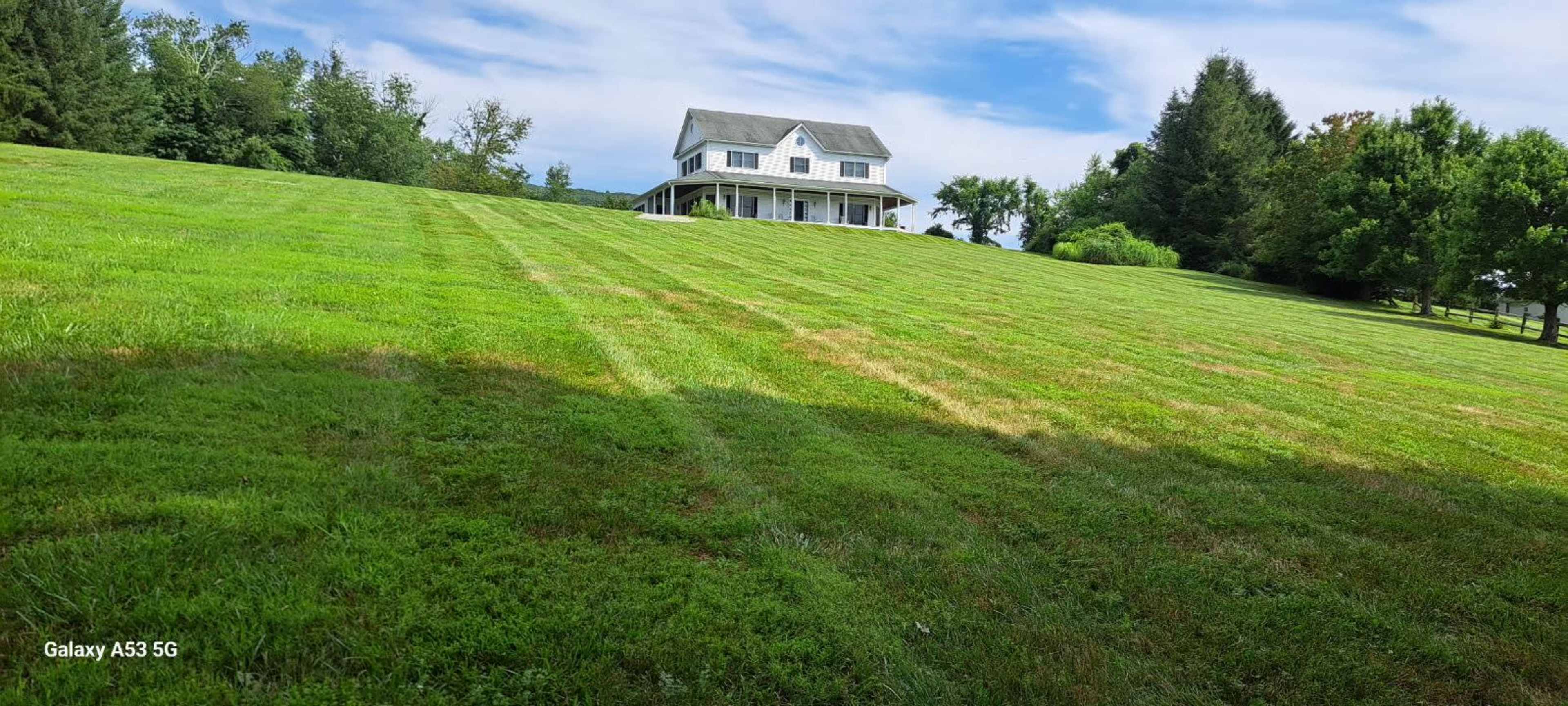 A large house with a front porch sits atop a sloped green lawn with neatly mowed grass and trees in the background.