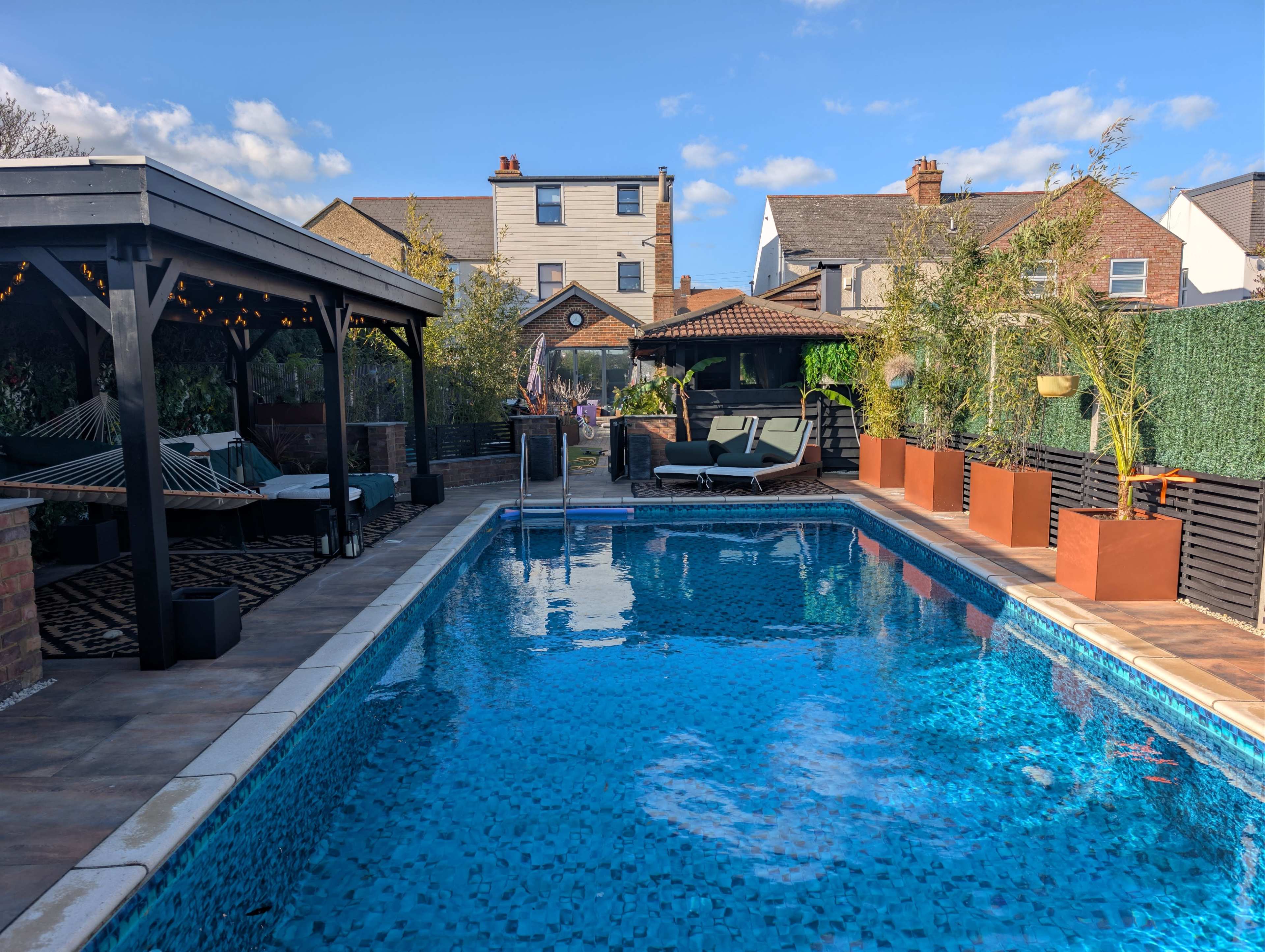 The image shows a residential backyard featuring a swimming pool surrounded by a wooden deck, lounge chairs, and potted plants against a backdrop of nearby houses.