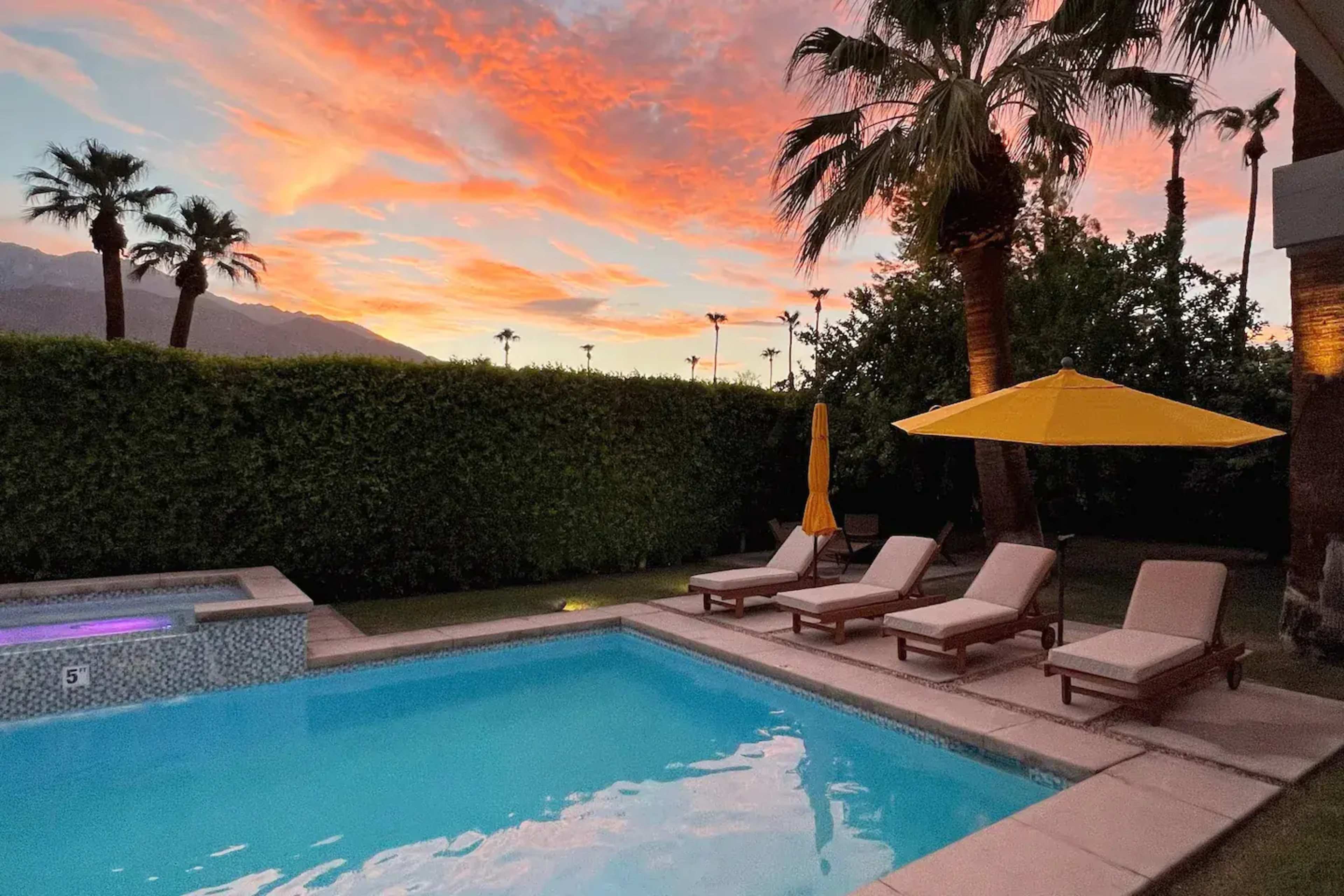 The image shows a swimming pool with lounge chairs and a yellow umbrella, set against a vibrant sunset sky and palm trees.