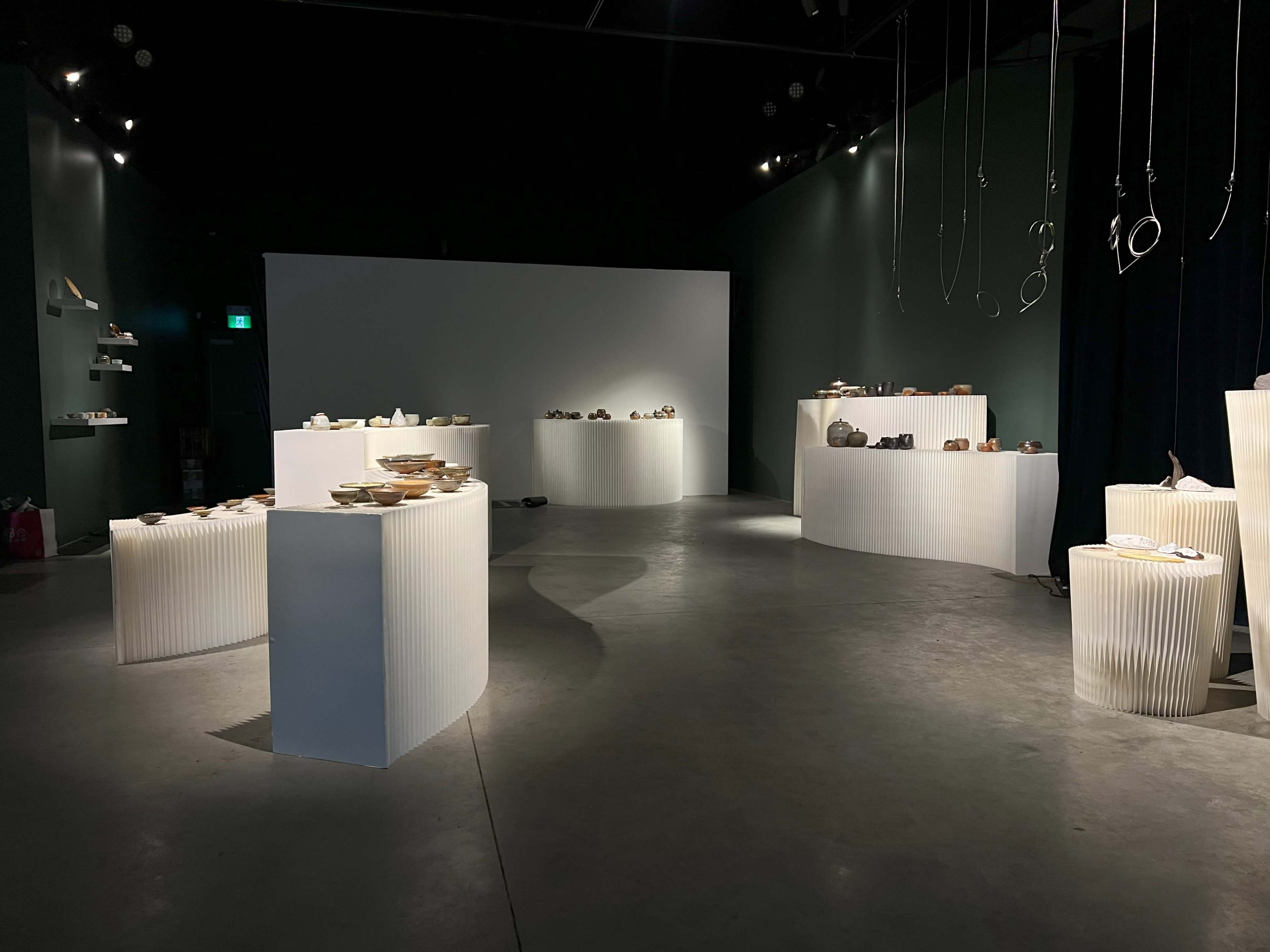 The image shows an exhibition space with several display platforms showcasing various ceramic pieces under dim lighting.
