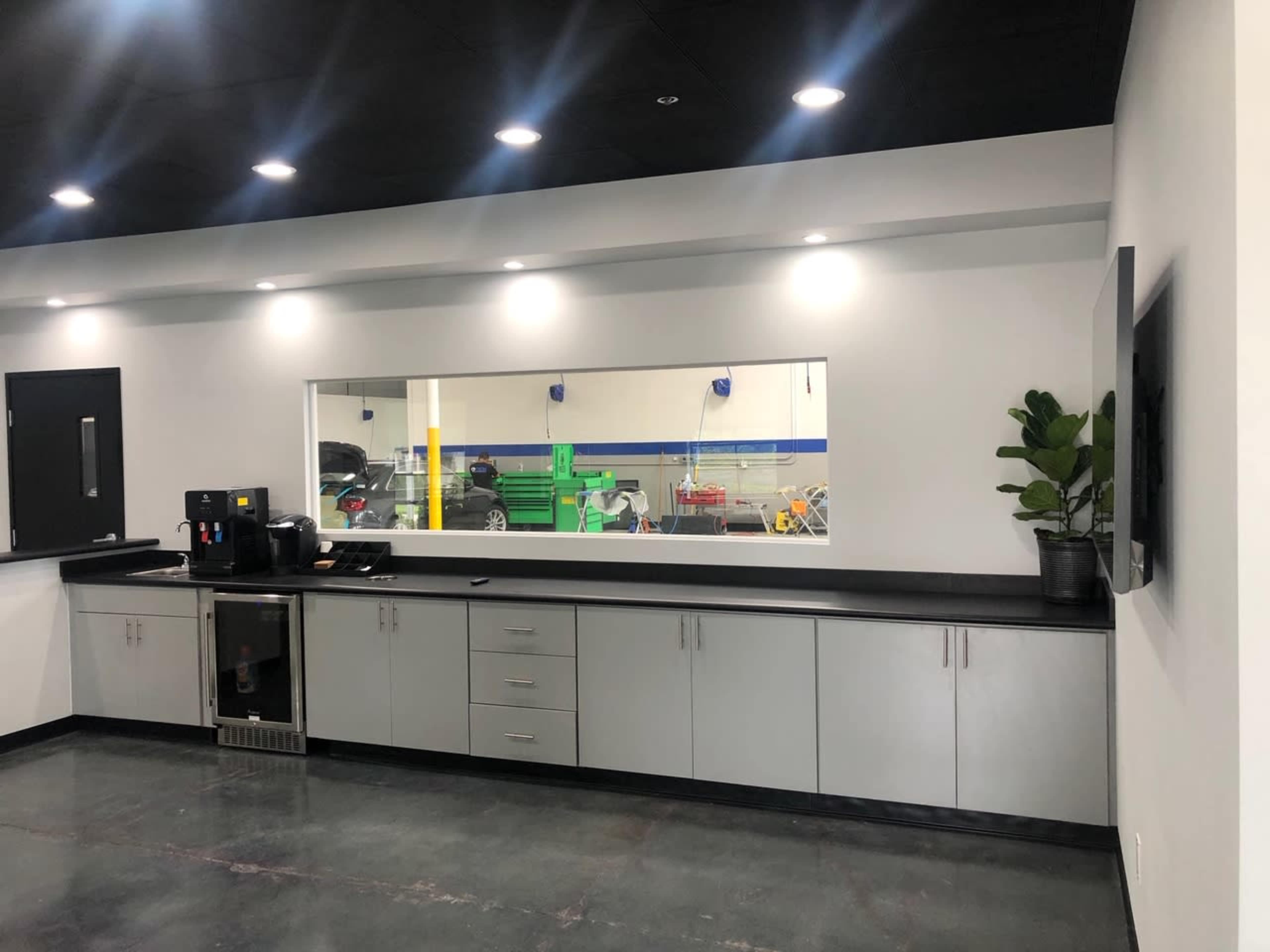 The image shows a modern kitchen area with a long countertop, a coffee machine, a small refrigerator, and a large window revealing a workspace behind it.