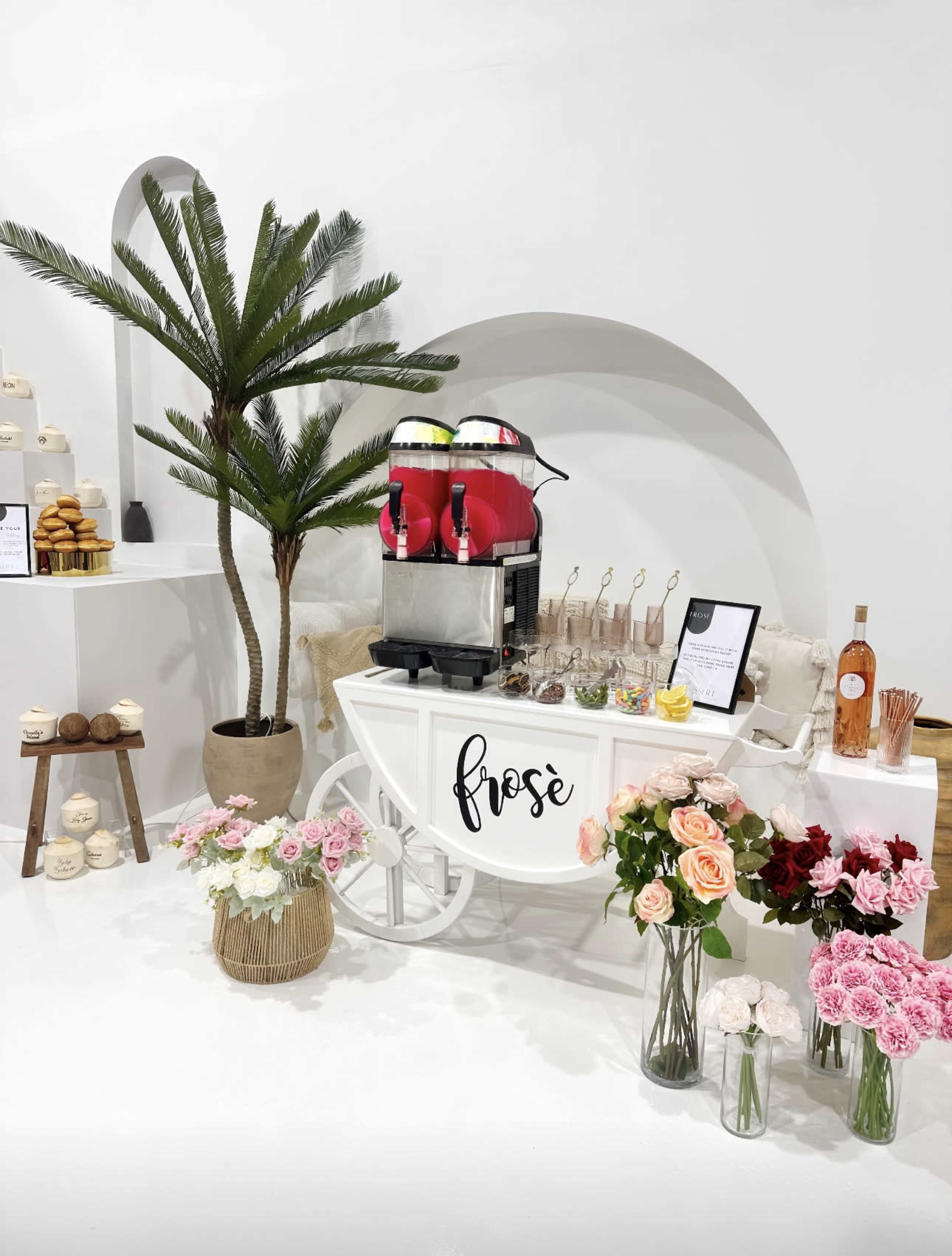 A white cart labeled "frosé" is set up with drink dispensers and surrounded by palm leaves, flowers, and sweets.