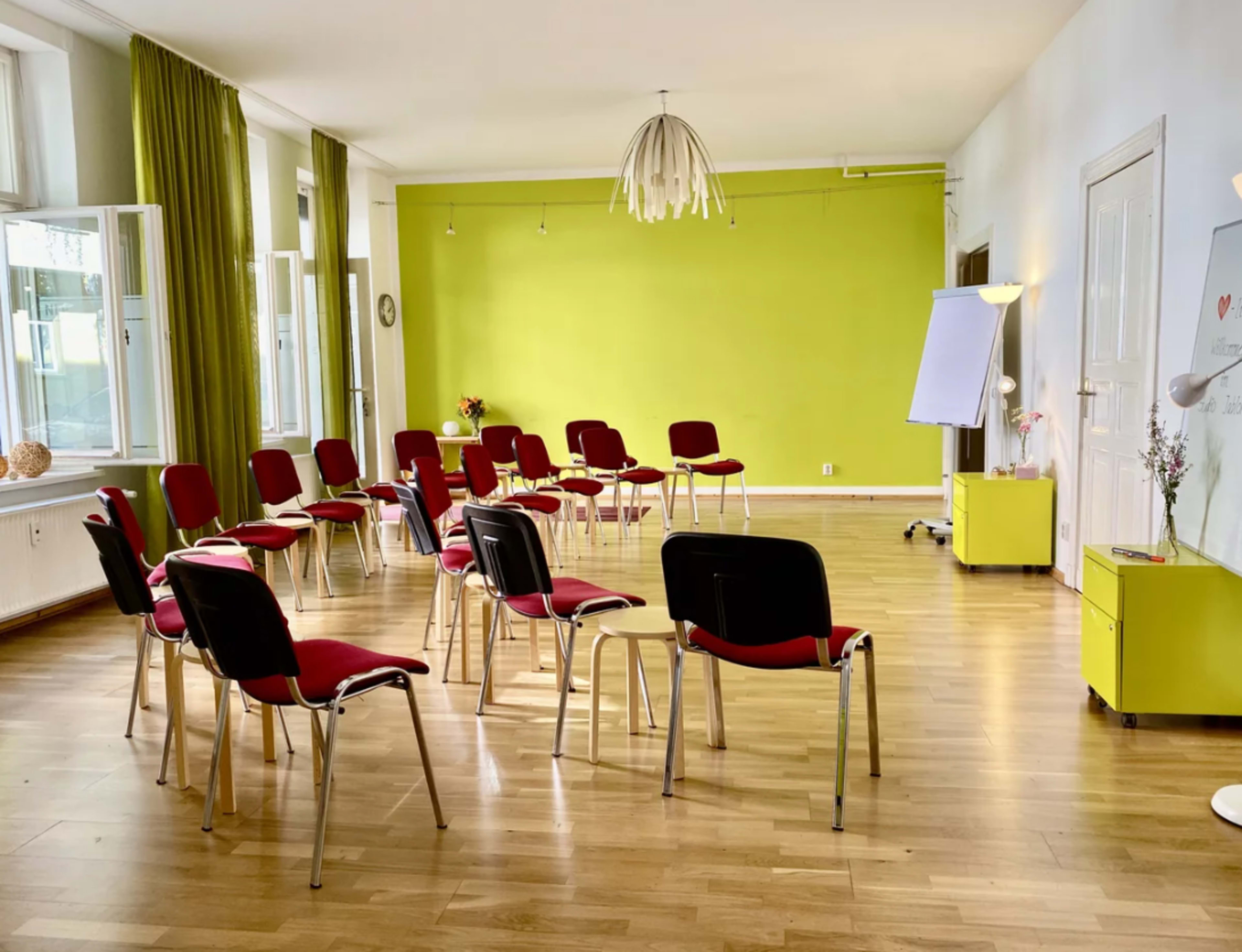 The image shows a room arranged with red chairs in a semi-circle, facing a blank presentation board against a bright green wall.