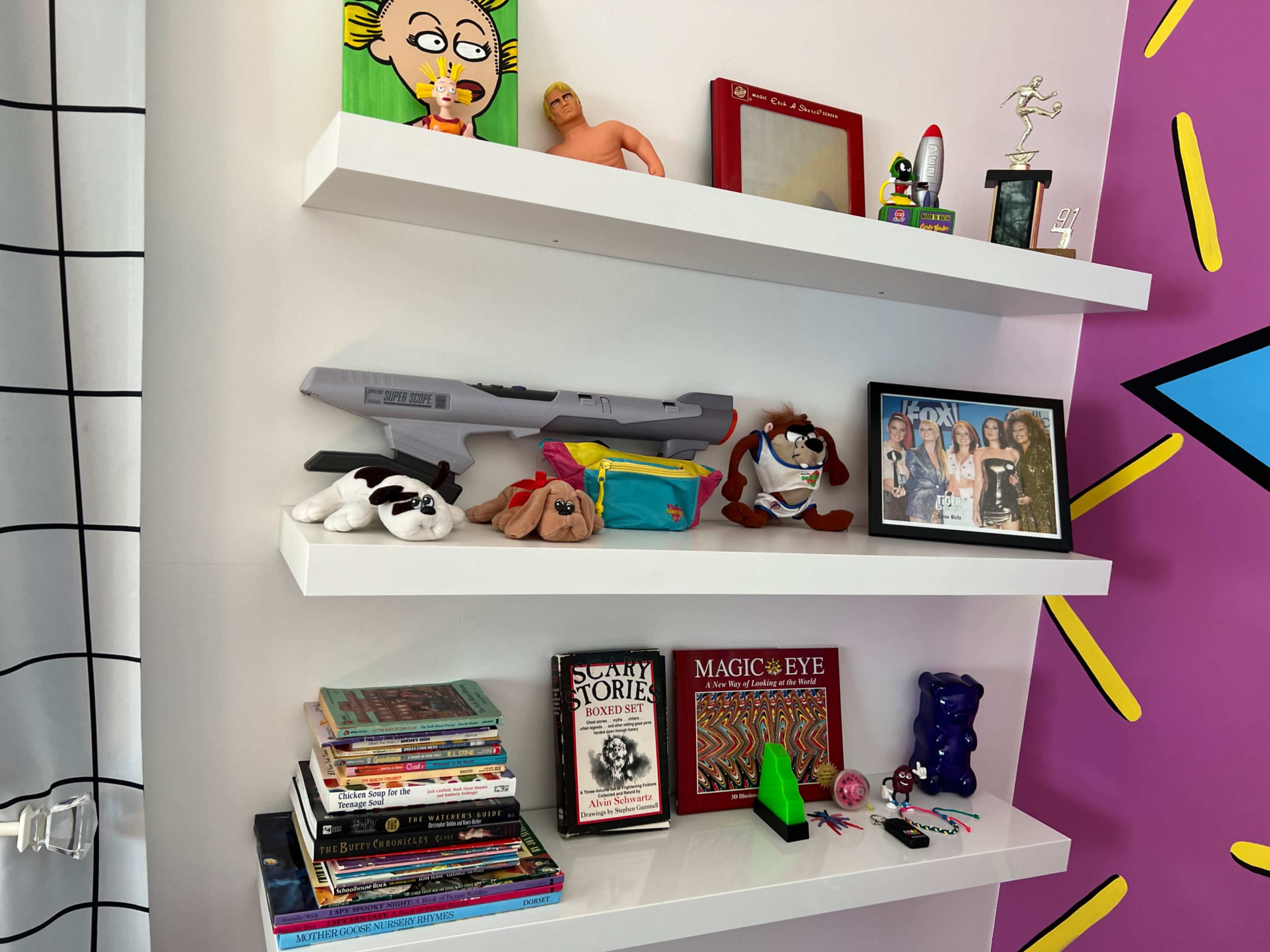 Three white shelves displaying various toys, books, a framed photo, and a trophy on a colorful wall background.