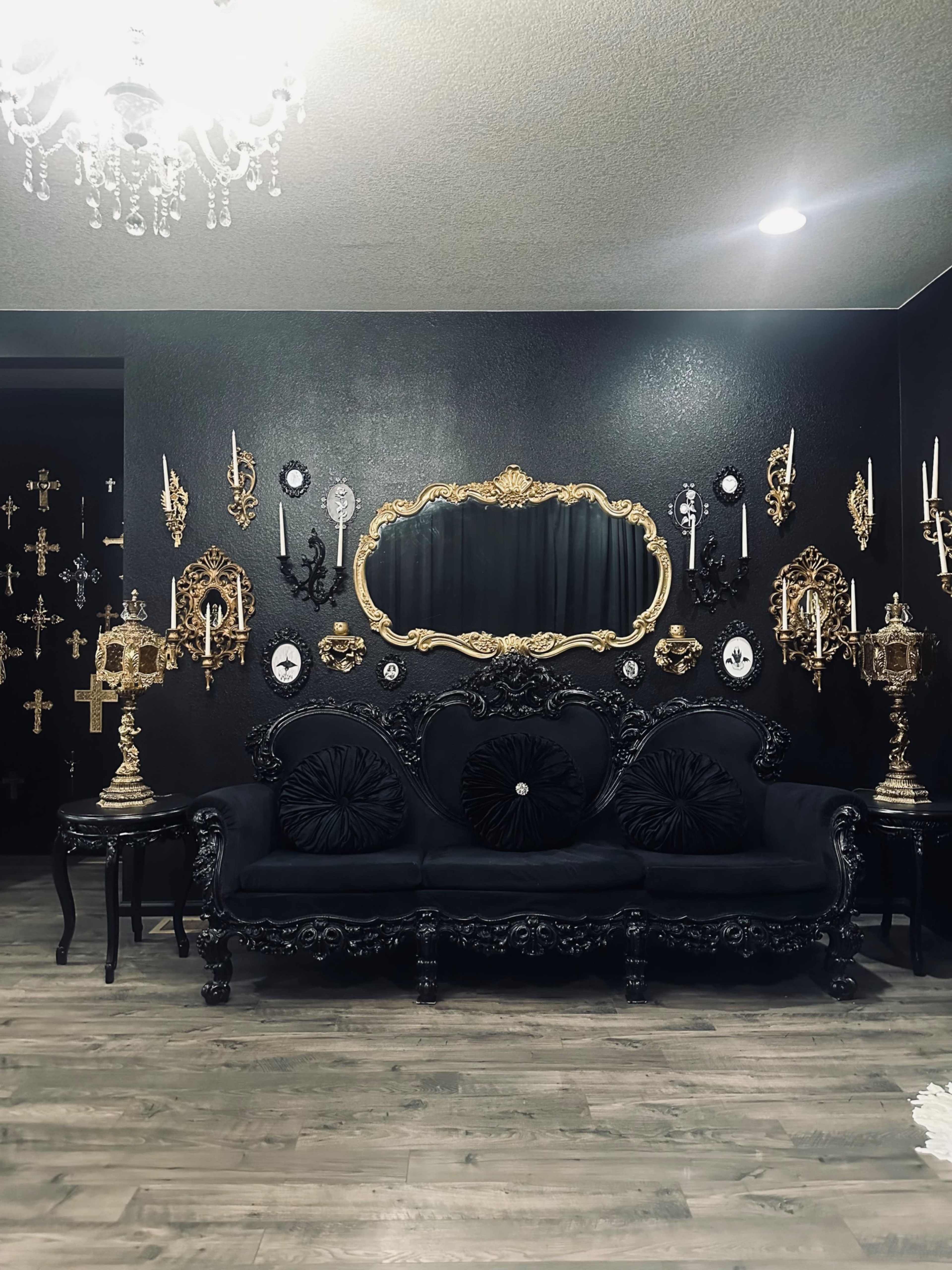 An elegantly adorned room featuring a black ornate couch, a large gilded mirror, and a collection of decorative wall sconces arranged on a dark wall.