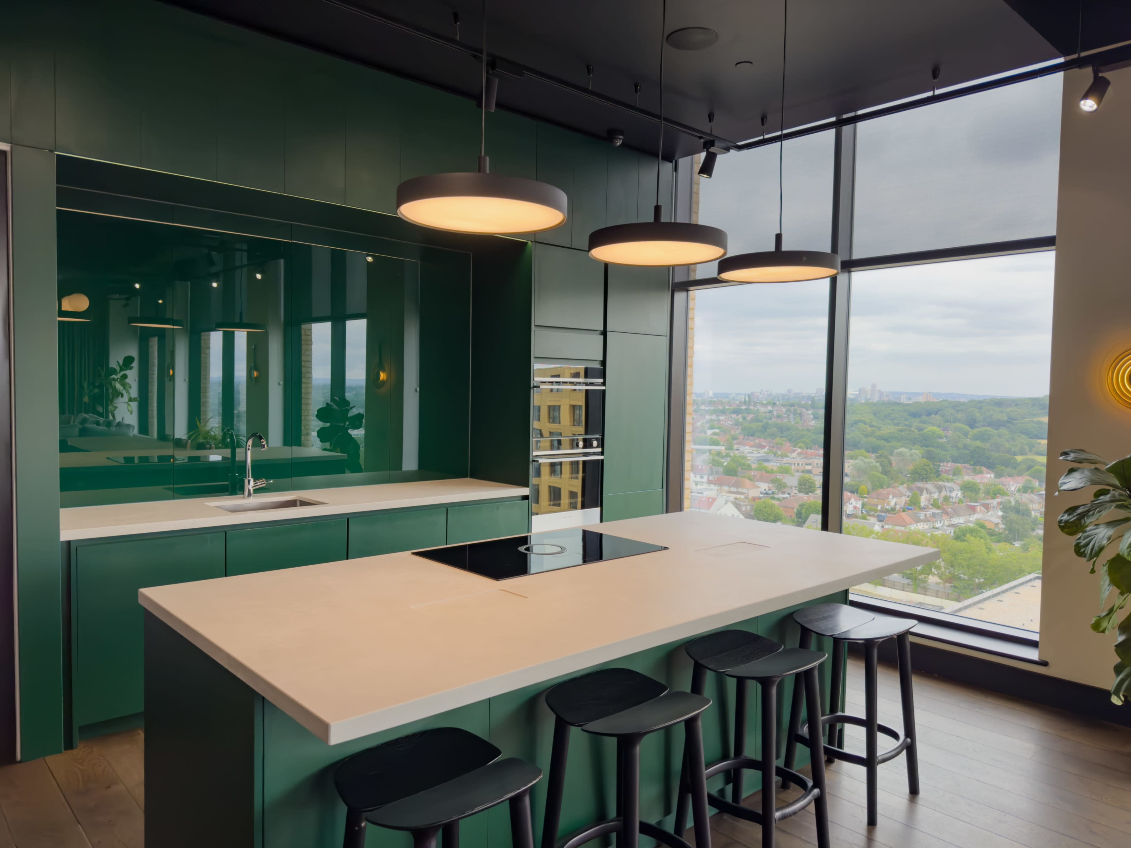 The image shows a modern kitchen with a green cabinetry and an island with seating, illuminated by pendant lights, overlooking a cityscape through large windows.