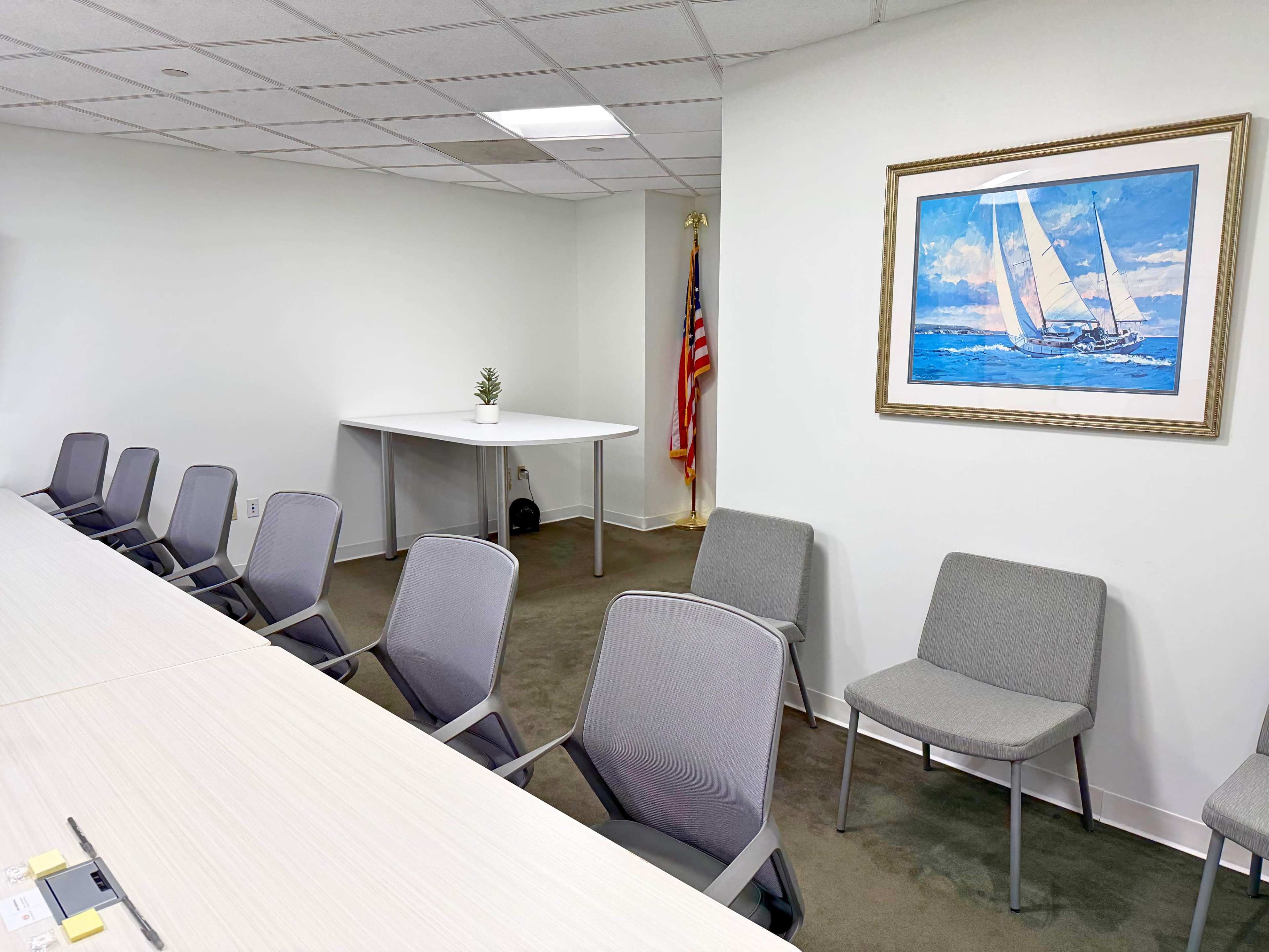 A conference room is set up with a long table, several gray chairs, a small table in the corner, and a framed painting of a sailboat on the wall, alongside an American flag.