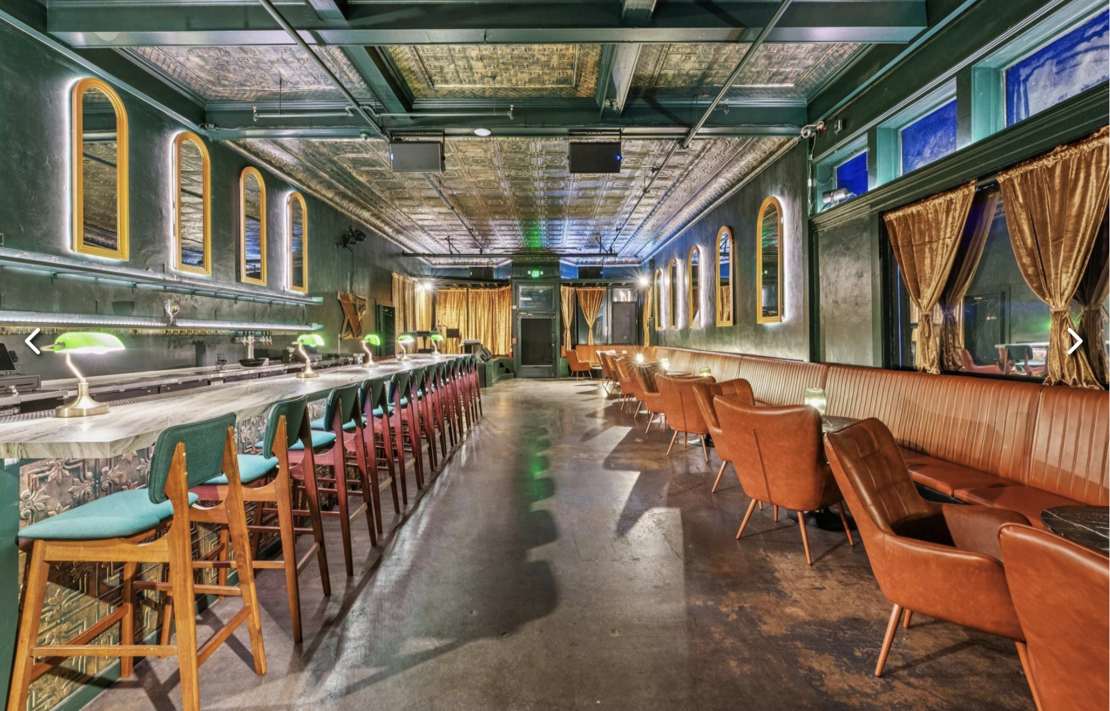 The image shows a stylish bar interior featuring a long counter with colorful stools, comfortable seating along the walls, and decorative lighting.