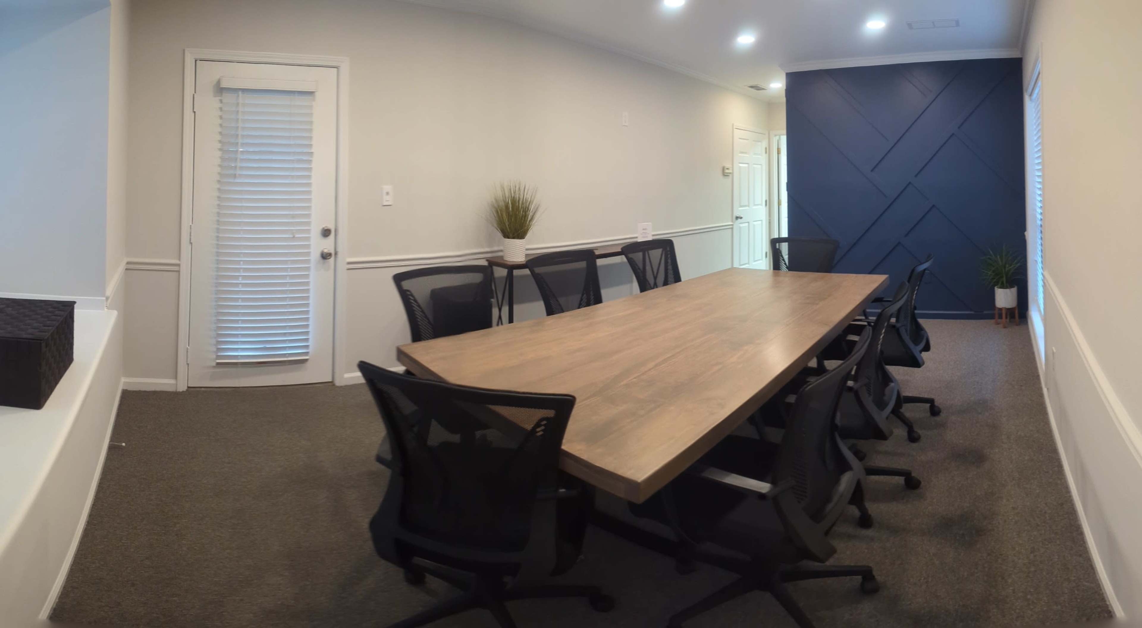 A spacious conference room features a long wooden table surrounded by black mesh chairs, with a door leading outside and a potted plant in the corner.