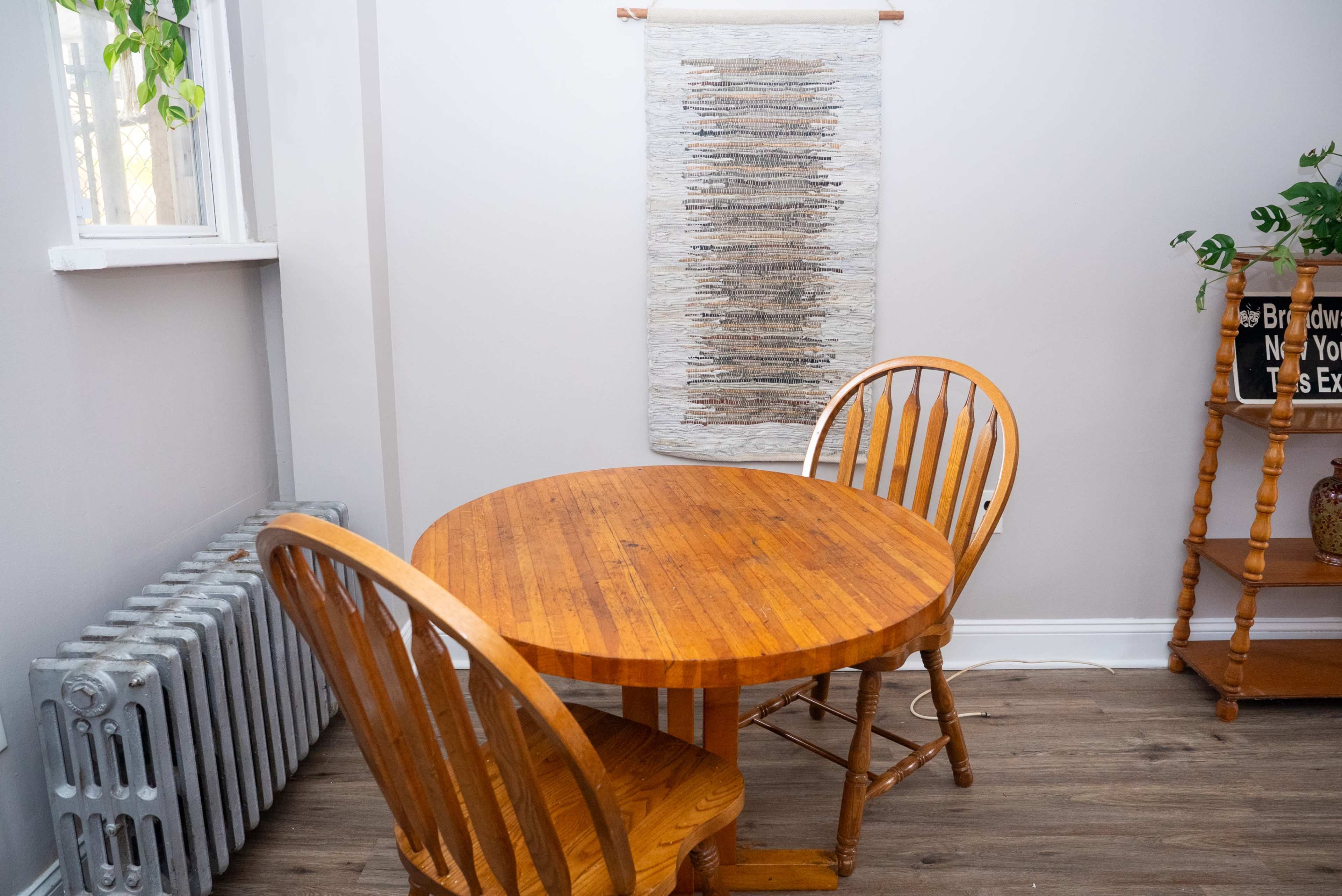 A round wooden table is set between two wooden chairs in a room featuring a radiator and a textured wall hanging.
