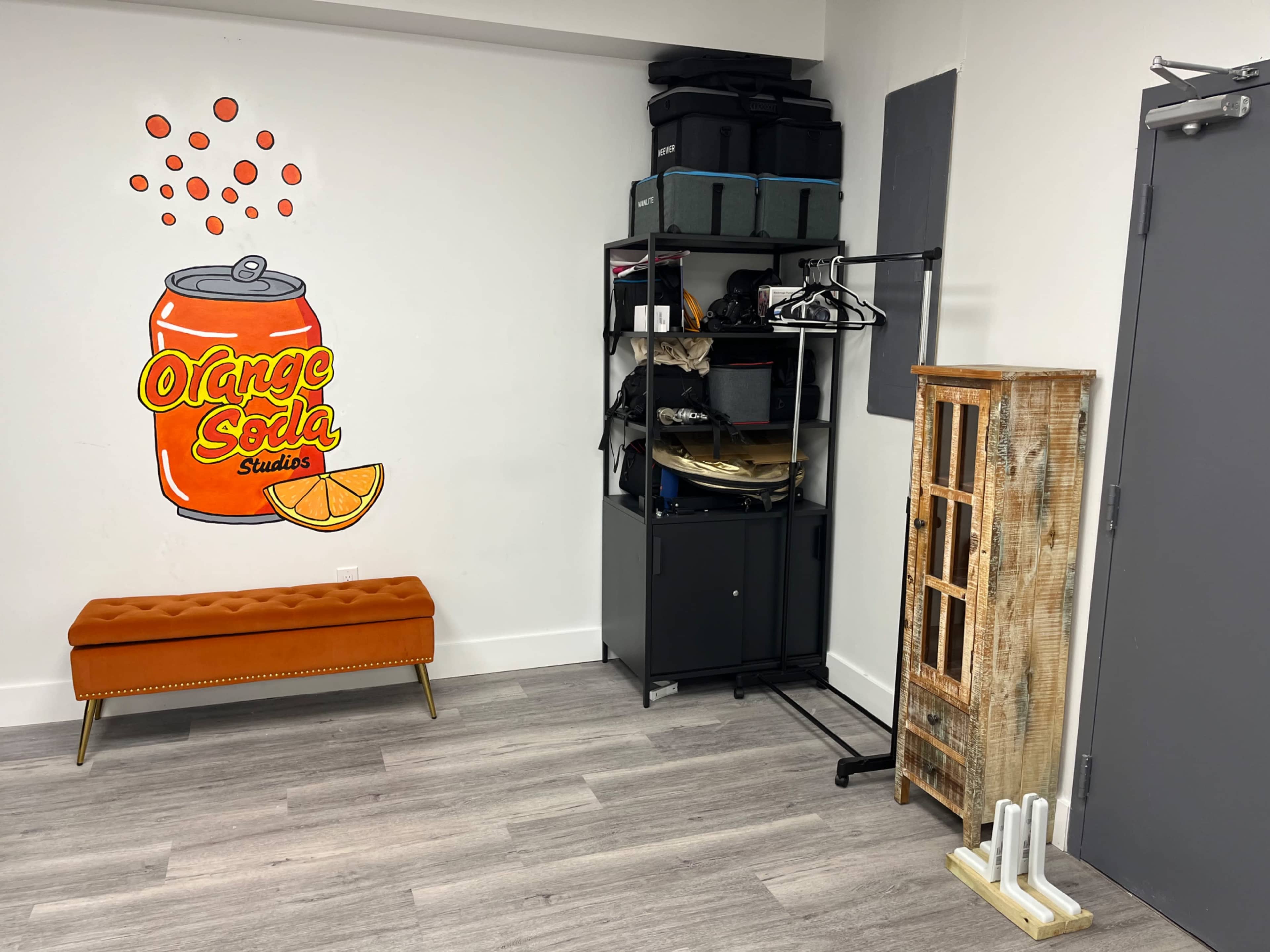 The image shows a room with a bright orange soda mural on the wall, a decorative orange bench, a metal storage rack filled with items, and a wooden storage unit.