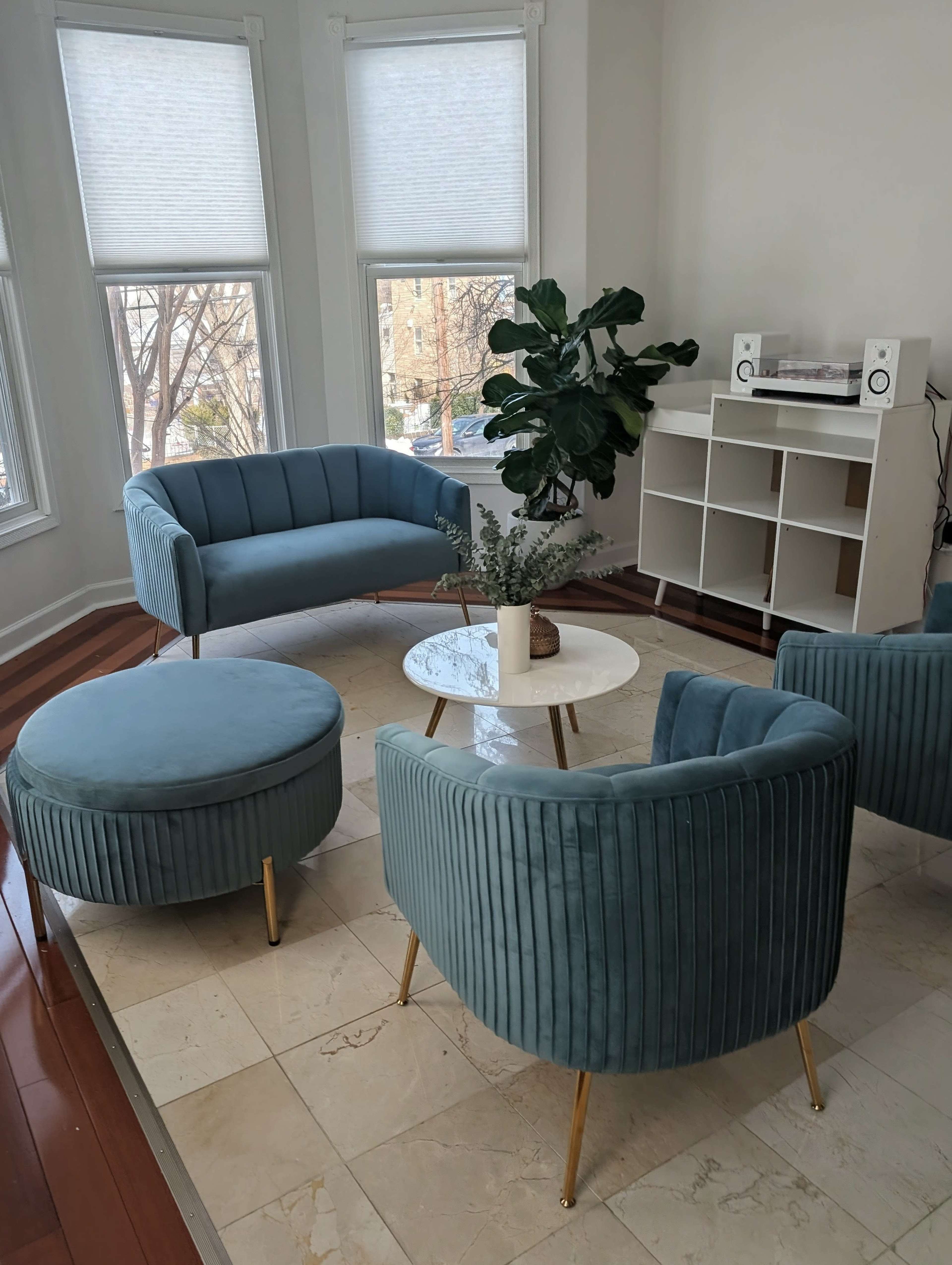 The image shows a modern living room with a light blue fabric sofa set, a round ottoman, a white coffee table, and large windows allowing natural light to enter.